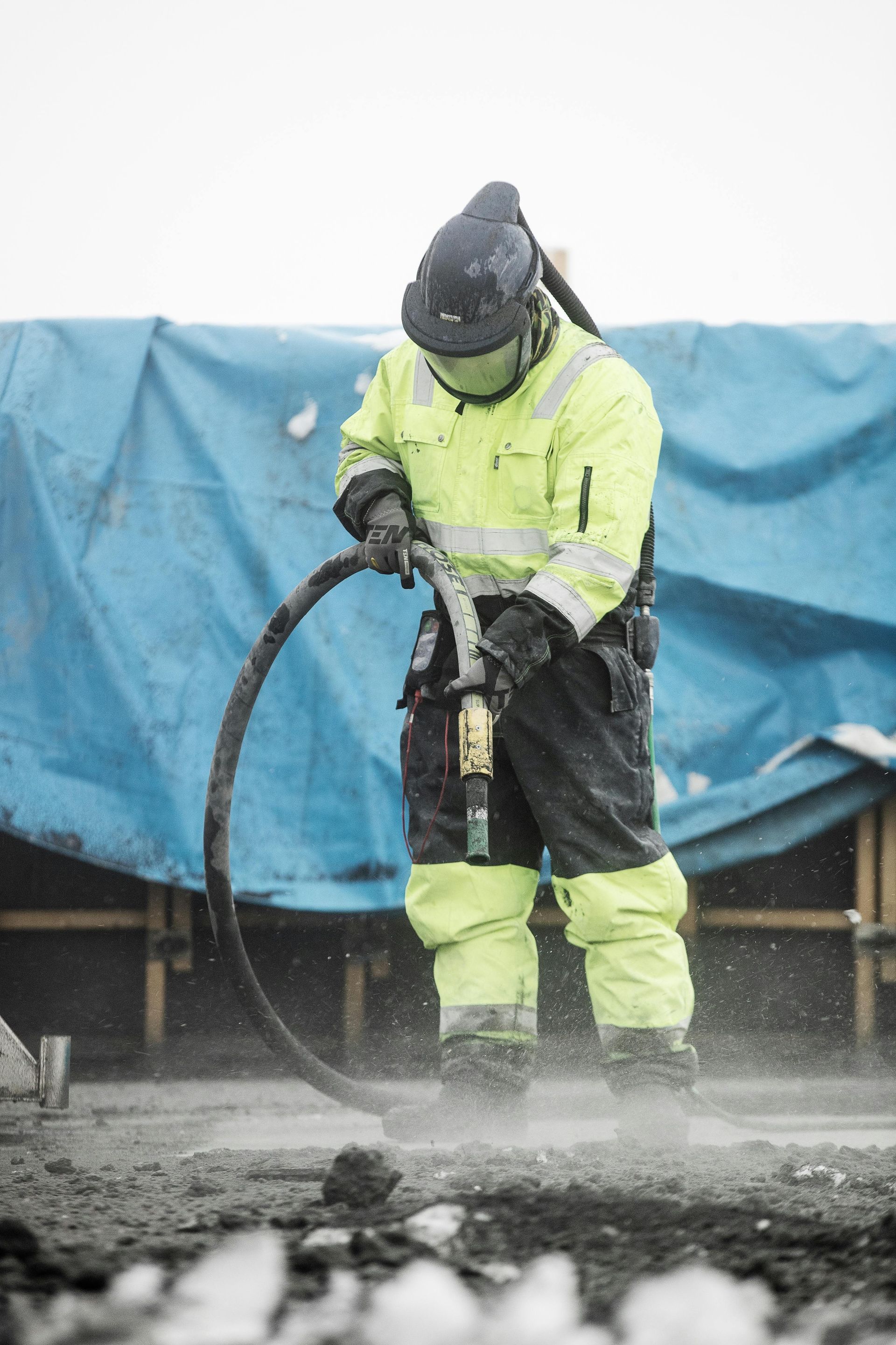 Construction worker in high-visibility gear using a jackhammer, kicking up dust.