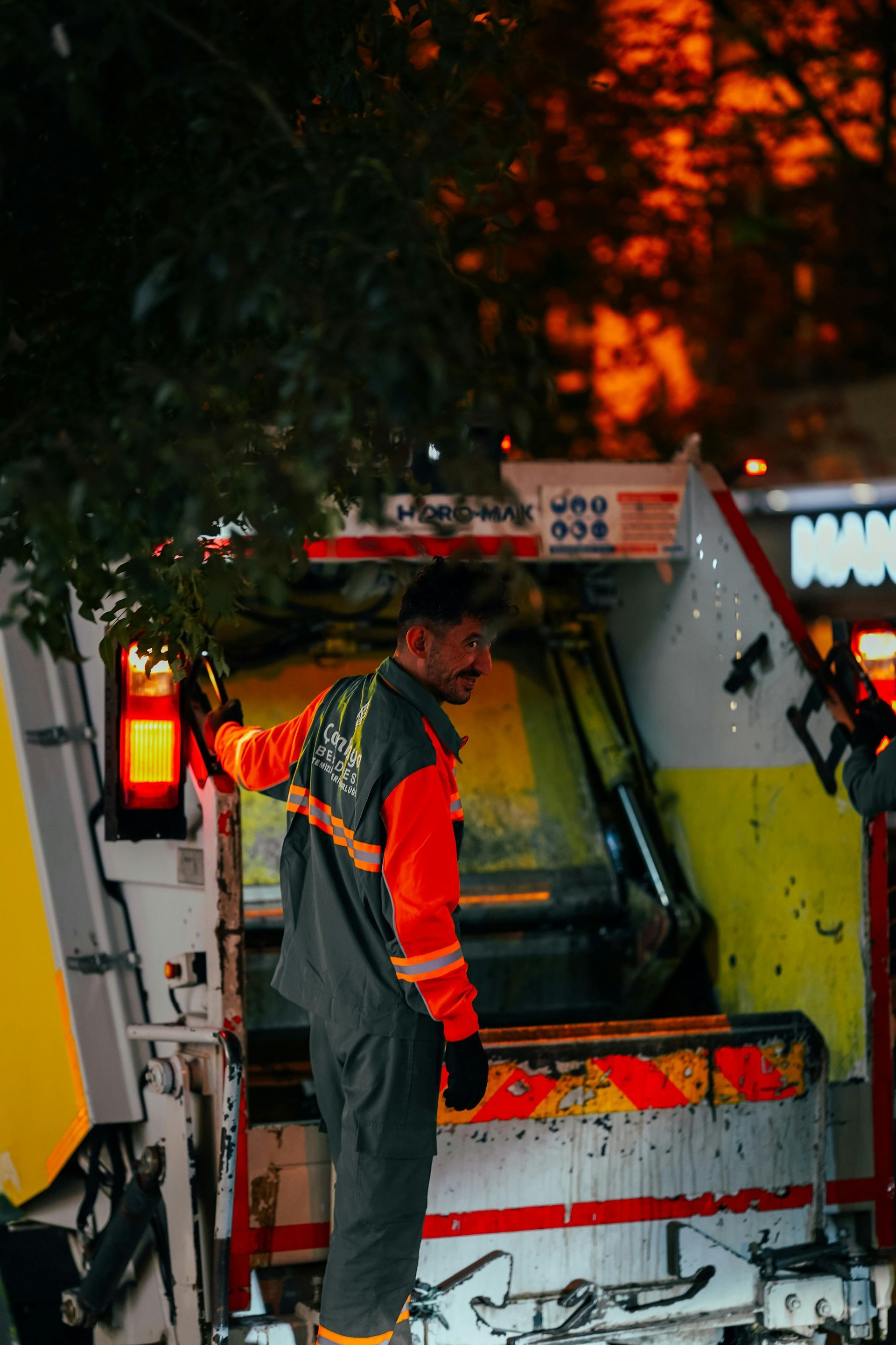 Garbage truck worker stands beside truck at night, wearing orange and gray uniform.
