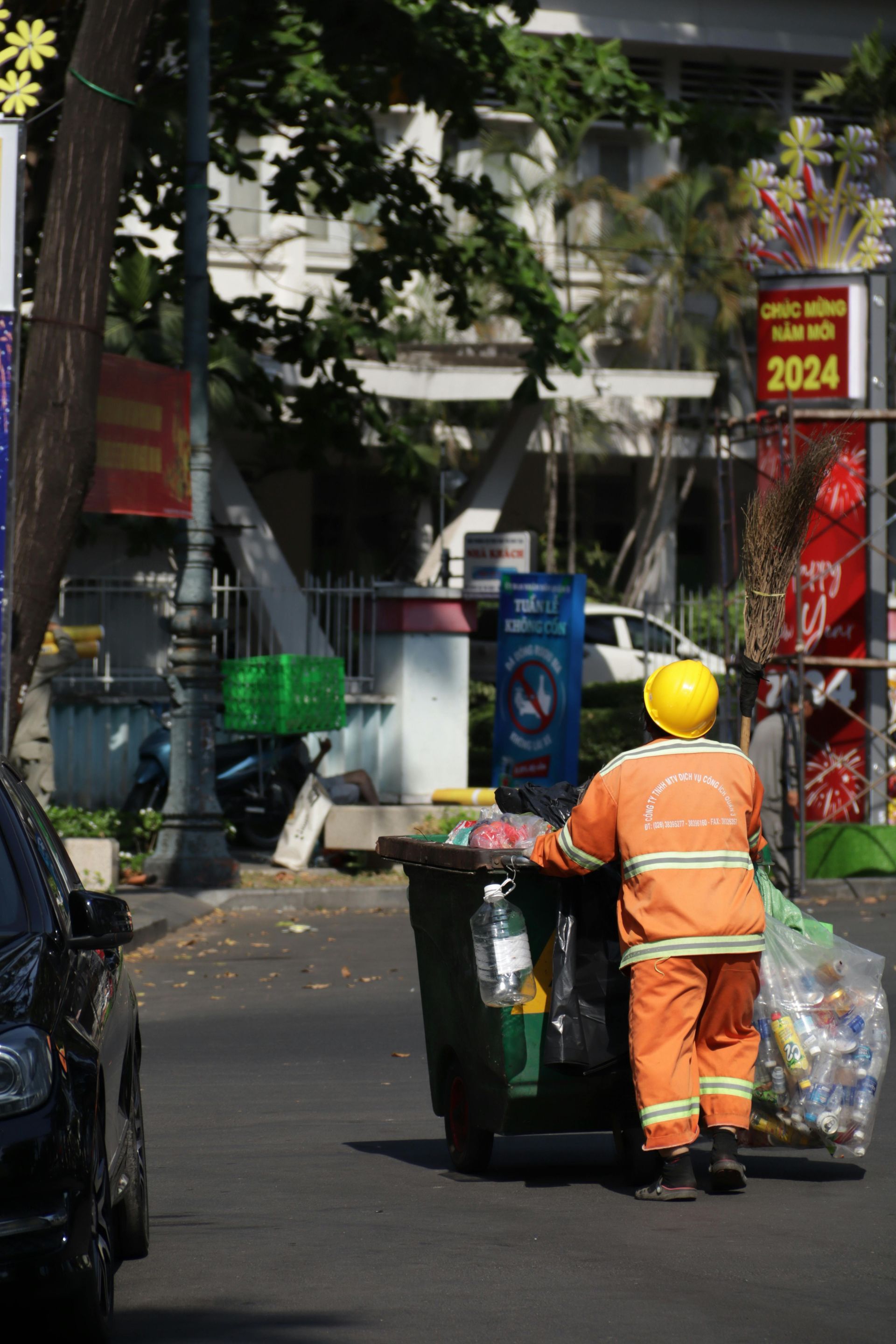 Street sweeper in orange uniform and yellow hard hat pushing a trash bin along a city street.