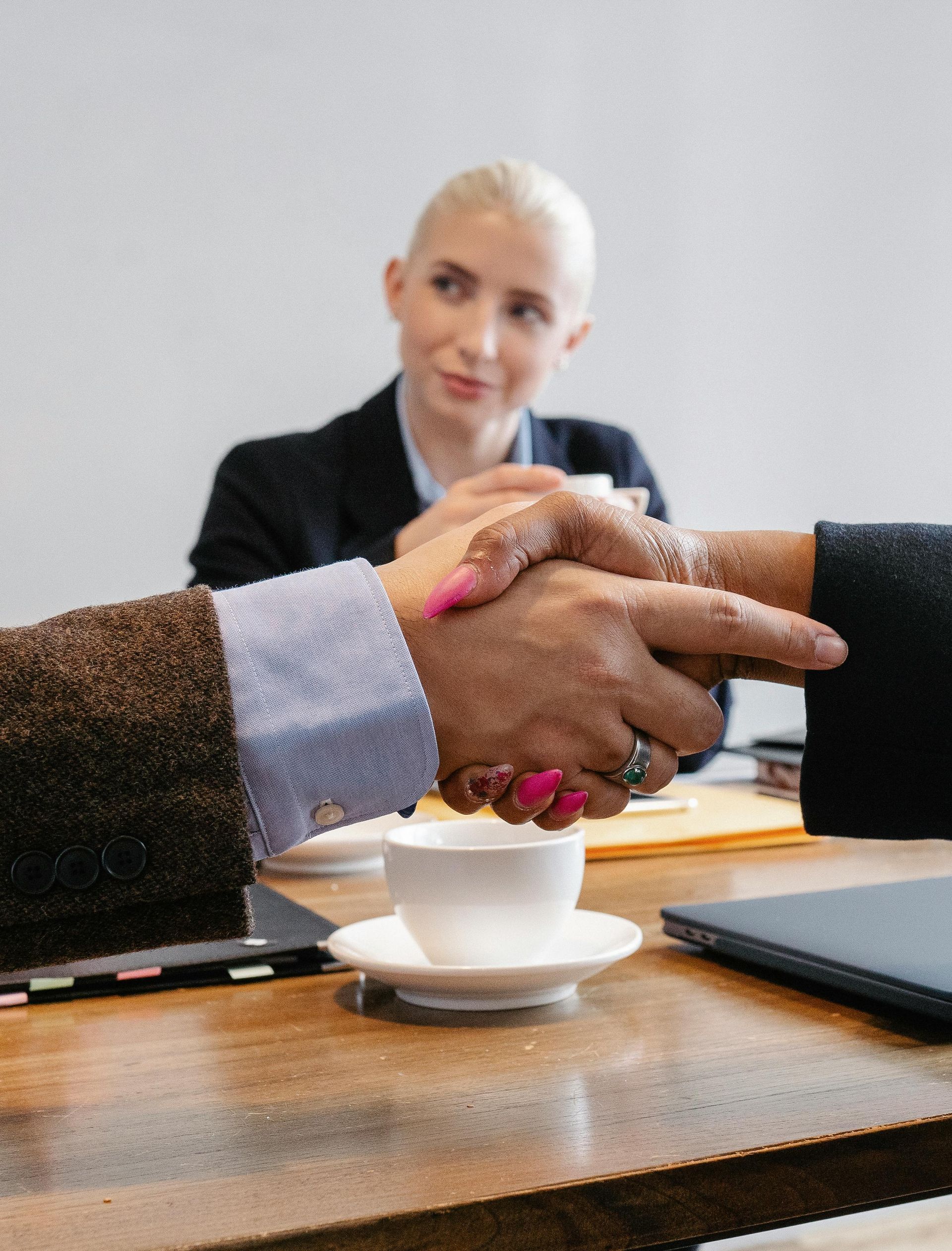 Two people shaking hands at a table, a woman in the background smiles.