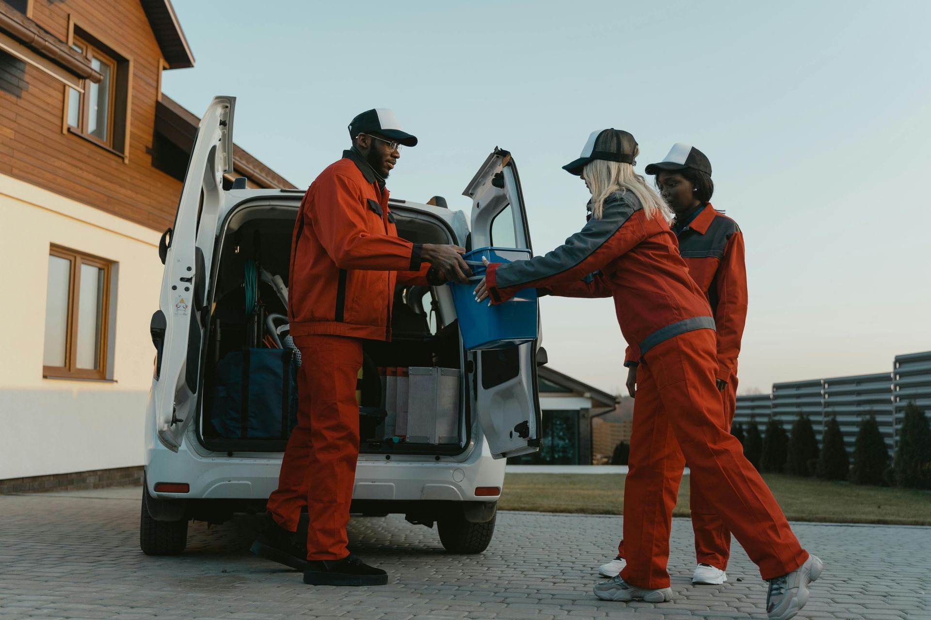 Five people in orange jumpsuits stand against a gray brick wall, leaning on blue trash bags.