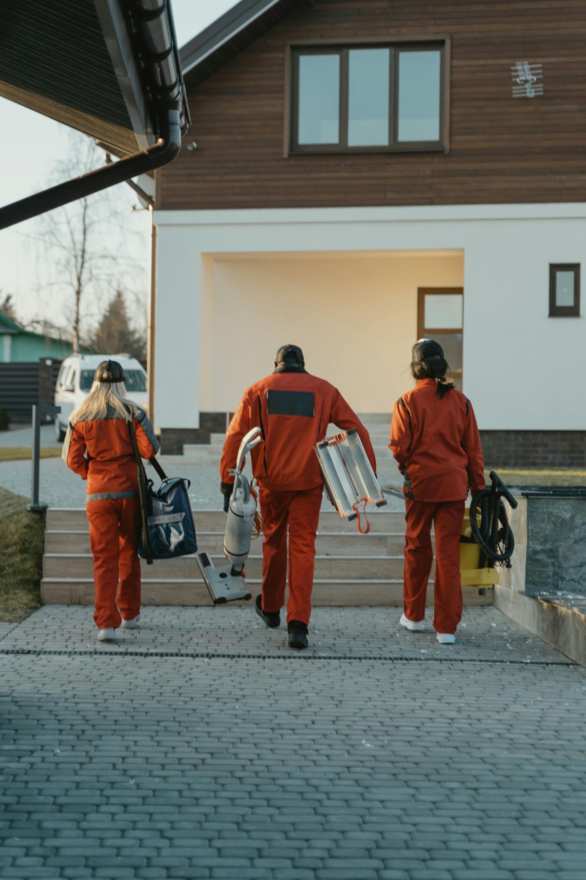 Three people in orange jumpsuits walk up steps towards a house, carrying tools and supplies.