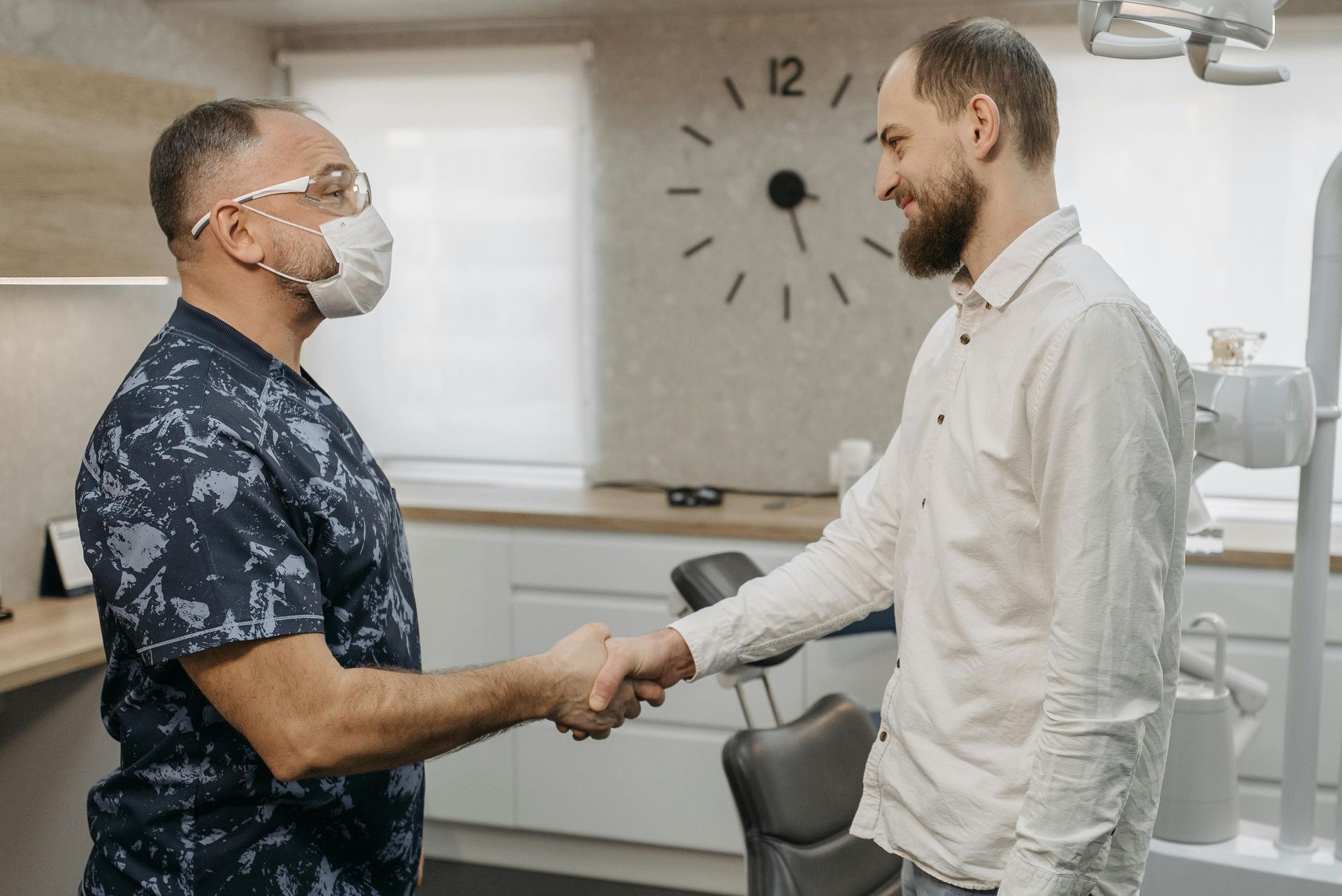 Dentist in scrubs and mask shakes hands with a patient in an office setting.
