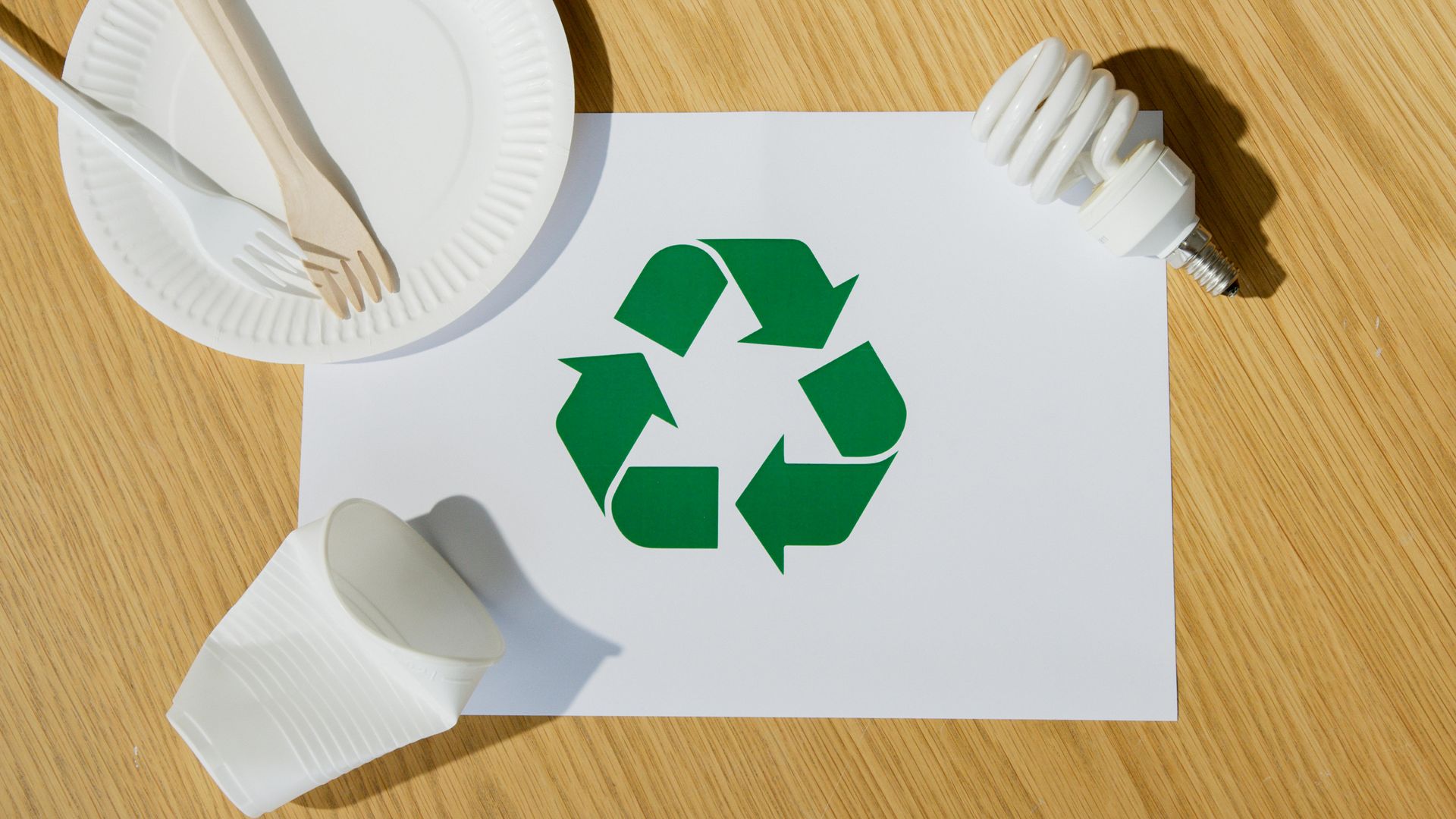 Recycling symbol on white paper with disposable plate, utensils, and lightbulb on wooden surface.