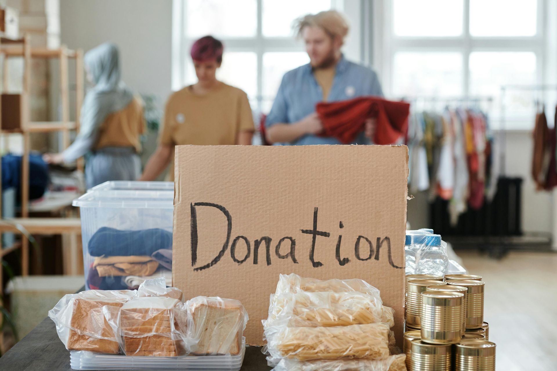 Donation box on table with food and supplies; volunteers sort items in background.