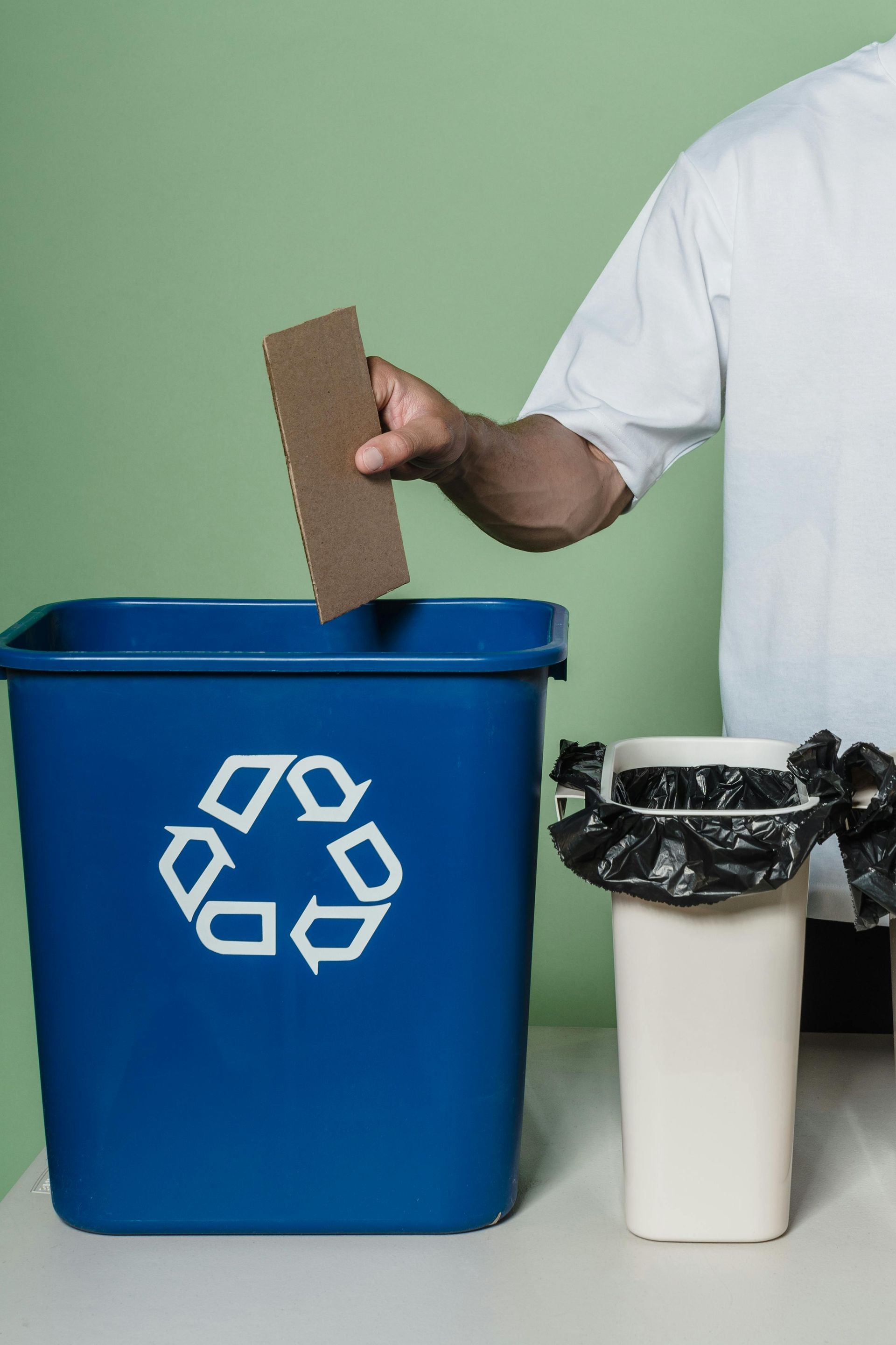 Person placing cardboard in a blue recycling bin. A white trash can sits beside it.