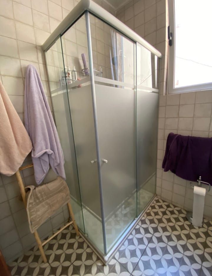 Mampara de ducha en baño con suelo de baldosas estampadas, toallas y silla.