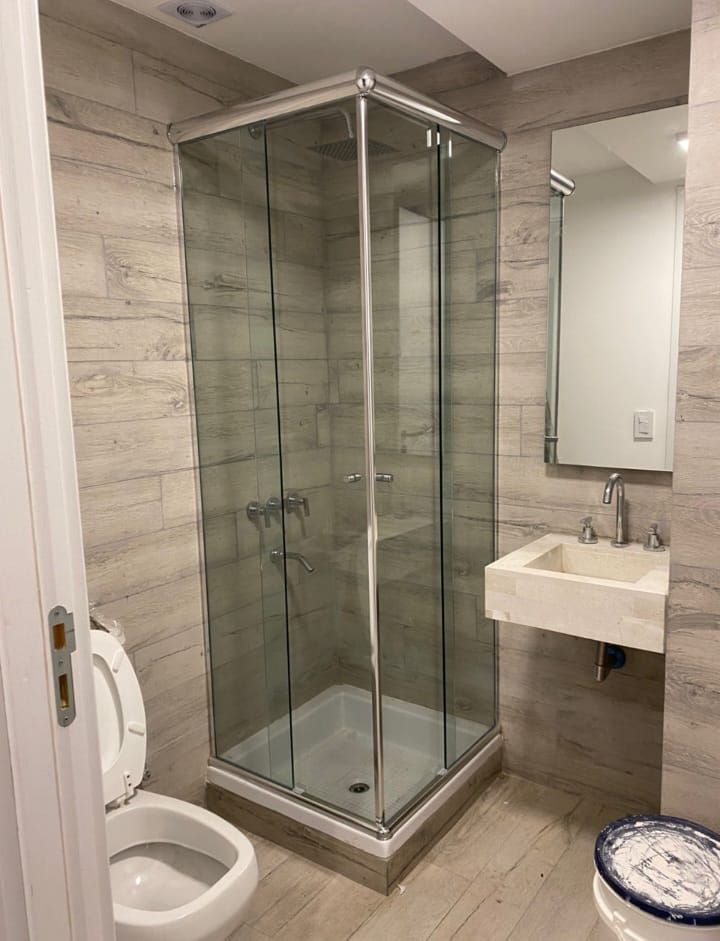 Baño con ducha de cristal, inodoro y lavabo con espejo. Paredes en tonos neutros.