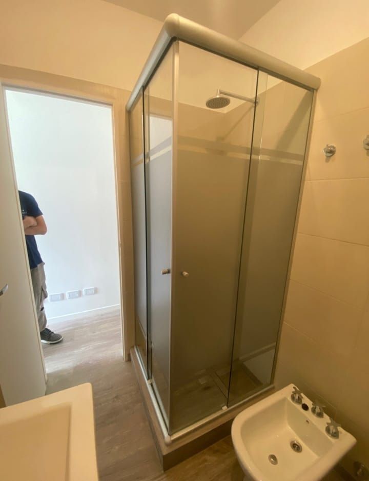 Cabina de ducha con puertas de cristal esmerilado en un baño. Una persona está de pie en una habitación contigua.