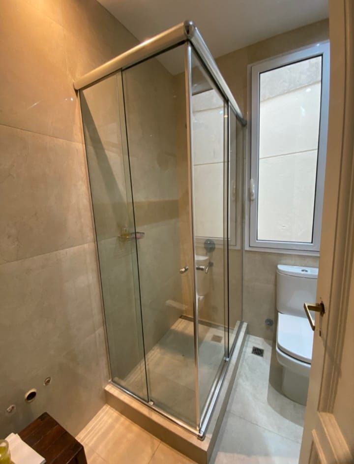 Mampara de ducha de cristal en baño con inodoro y ventana.