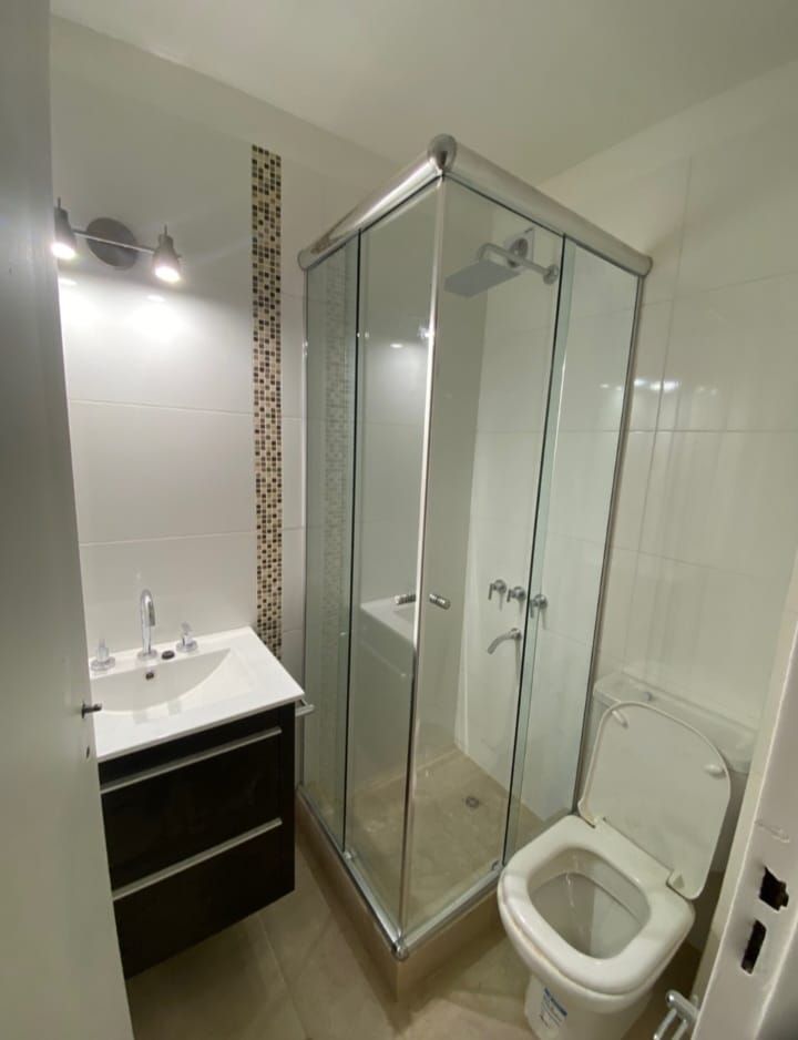 Baño con ducha de cristal, inodoro y lavabo con mueble oscuro. Paredes blancas y suelo color canela.