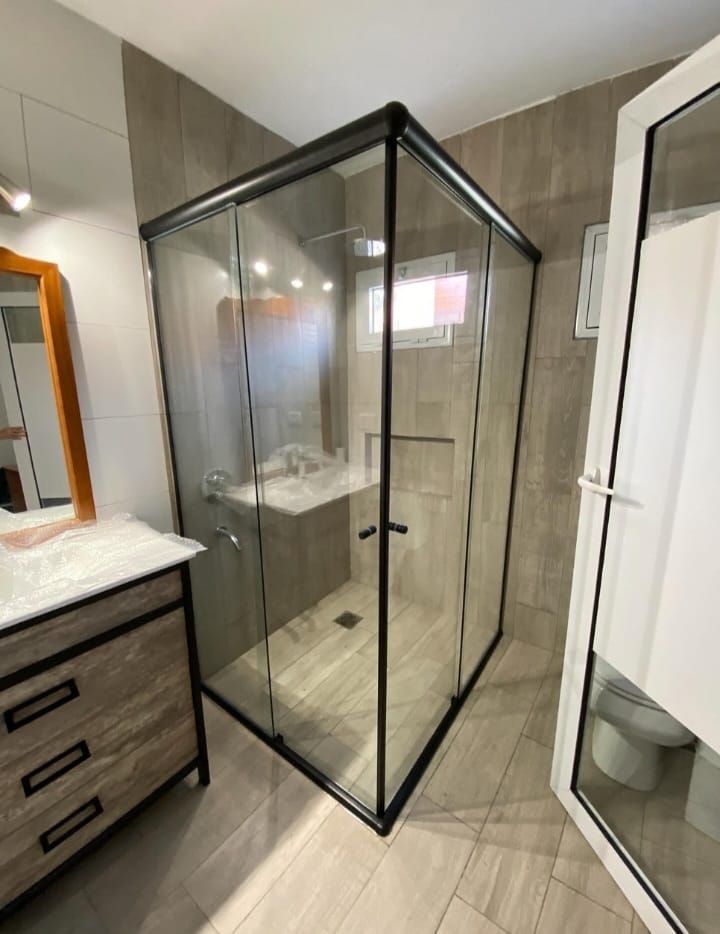 Baño moderno con mampara de ducha de cristal con marco negro. Tocador de madera clara y suelo de baldosas.