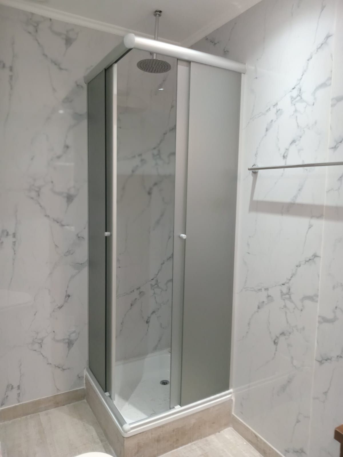 Cabina de ducha con puertas de cristal esmerilado y paredes de azulejos imitación mármol. Hay un toallero a la derecha.