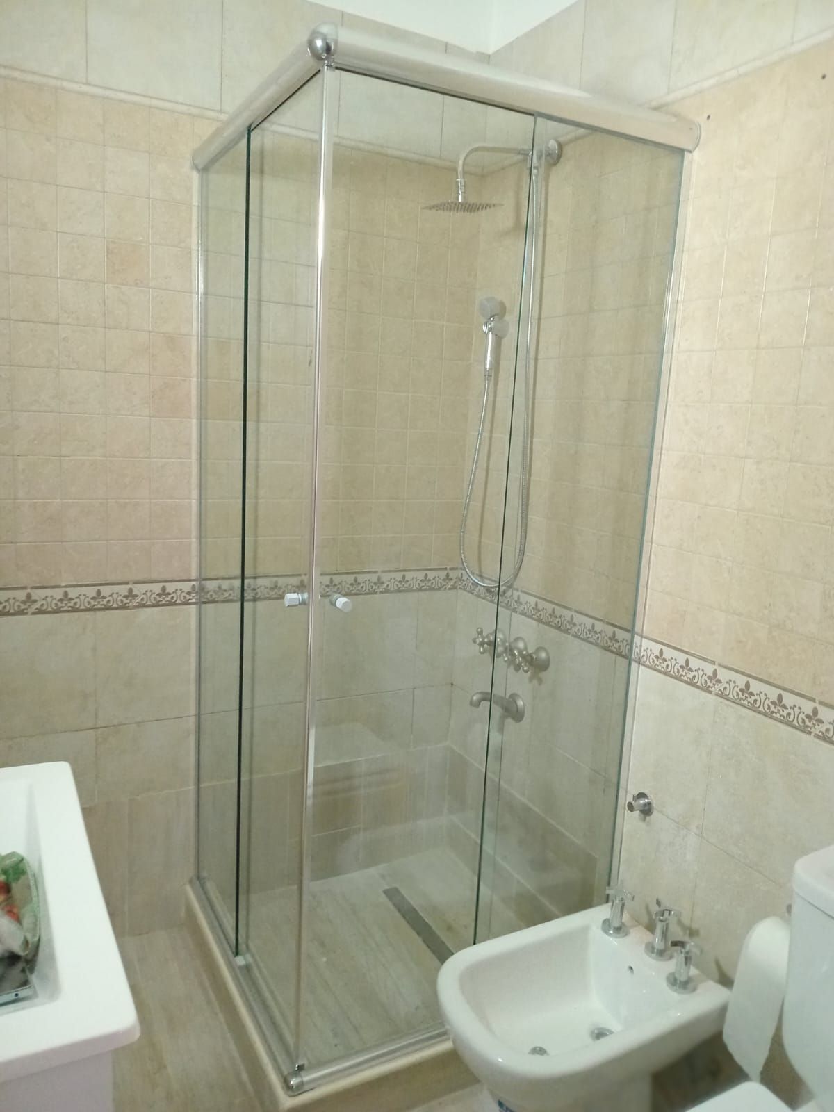 Cabina de ducha de cristal en un baño con paredes de azulejos de color beige y bidé.