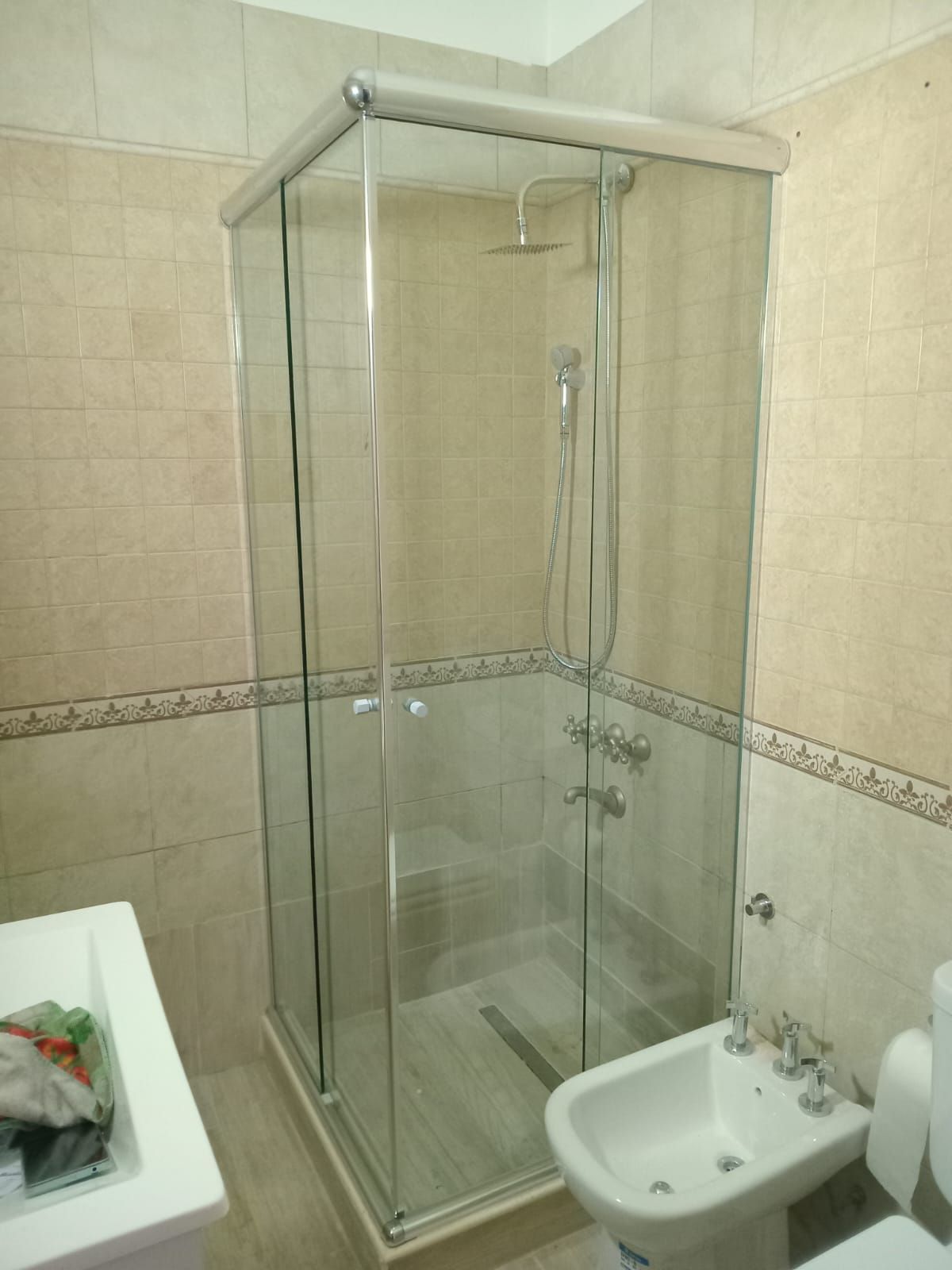 Baño con mampara de ducha de cristal, bidé y lavabo. Paredes de azulejos beige.