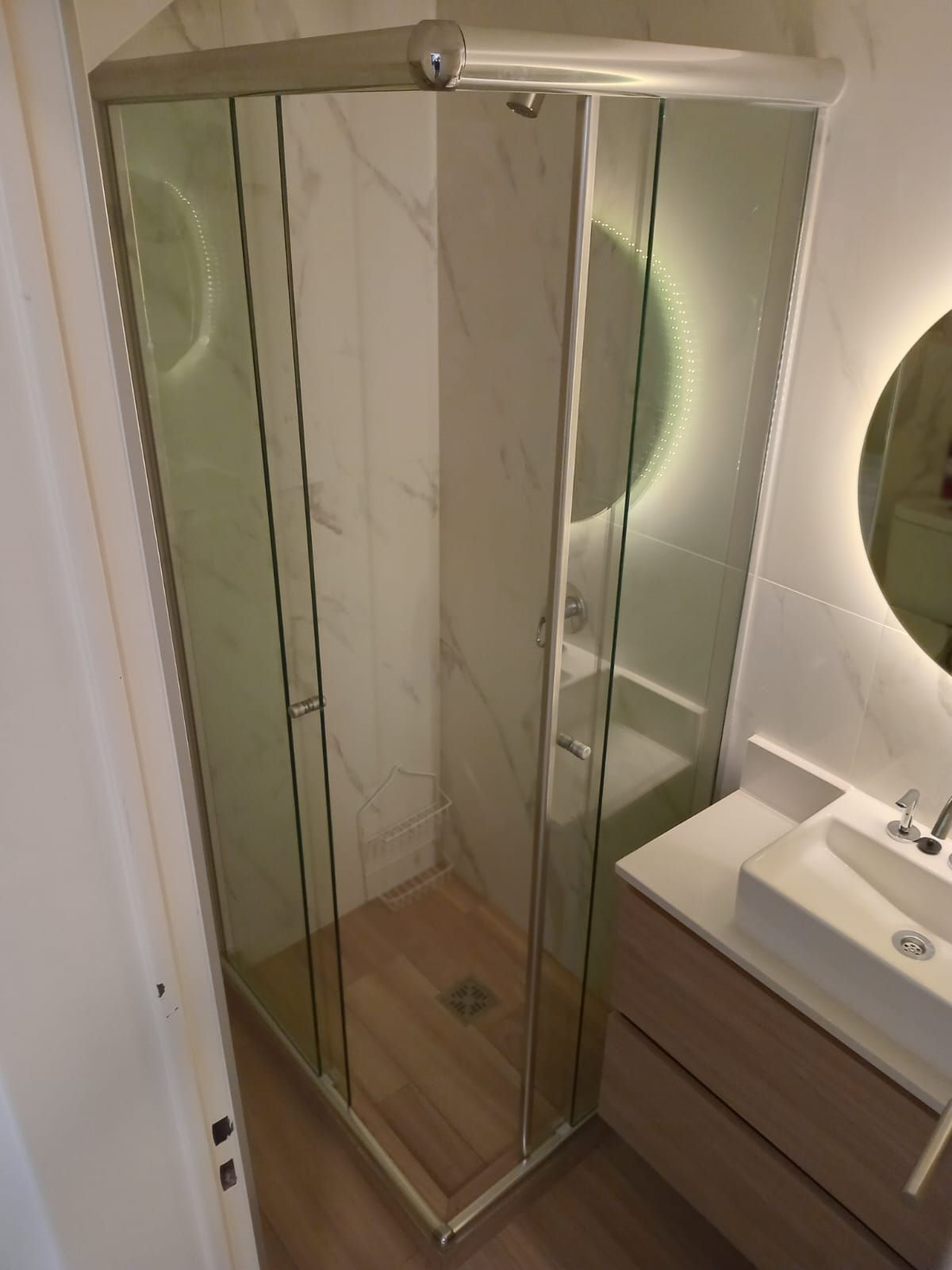 Cabina de ducha con puertas de cristal, paredes blancas, suelo símil madera, junto a un tocador con lavabo.