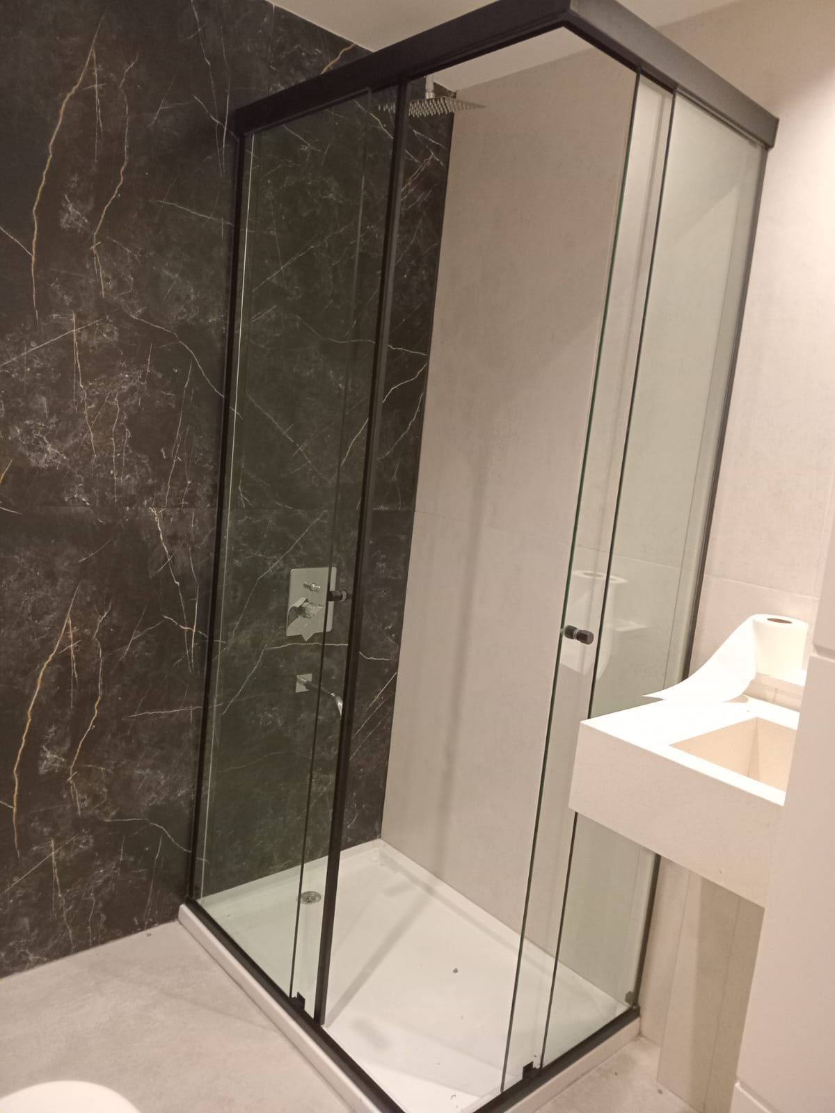 Mampara de ducha de cristal con marco negro, lavabo blanco y pared de mármol oscuro.