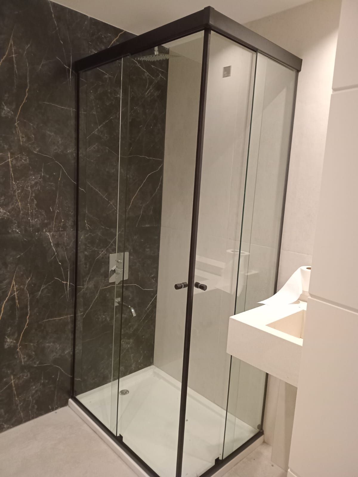 Mampara de ducha de cristal con marco negro y base de ducha de color blanco, junto a una pared con efecto mármol.