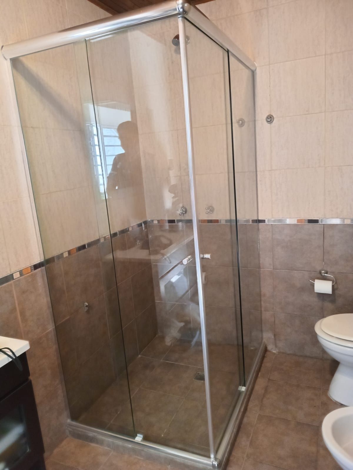 Cabina de ducha con puertas de cristal transparente y marco plateado en un baño con azulejos marrones y beige.