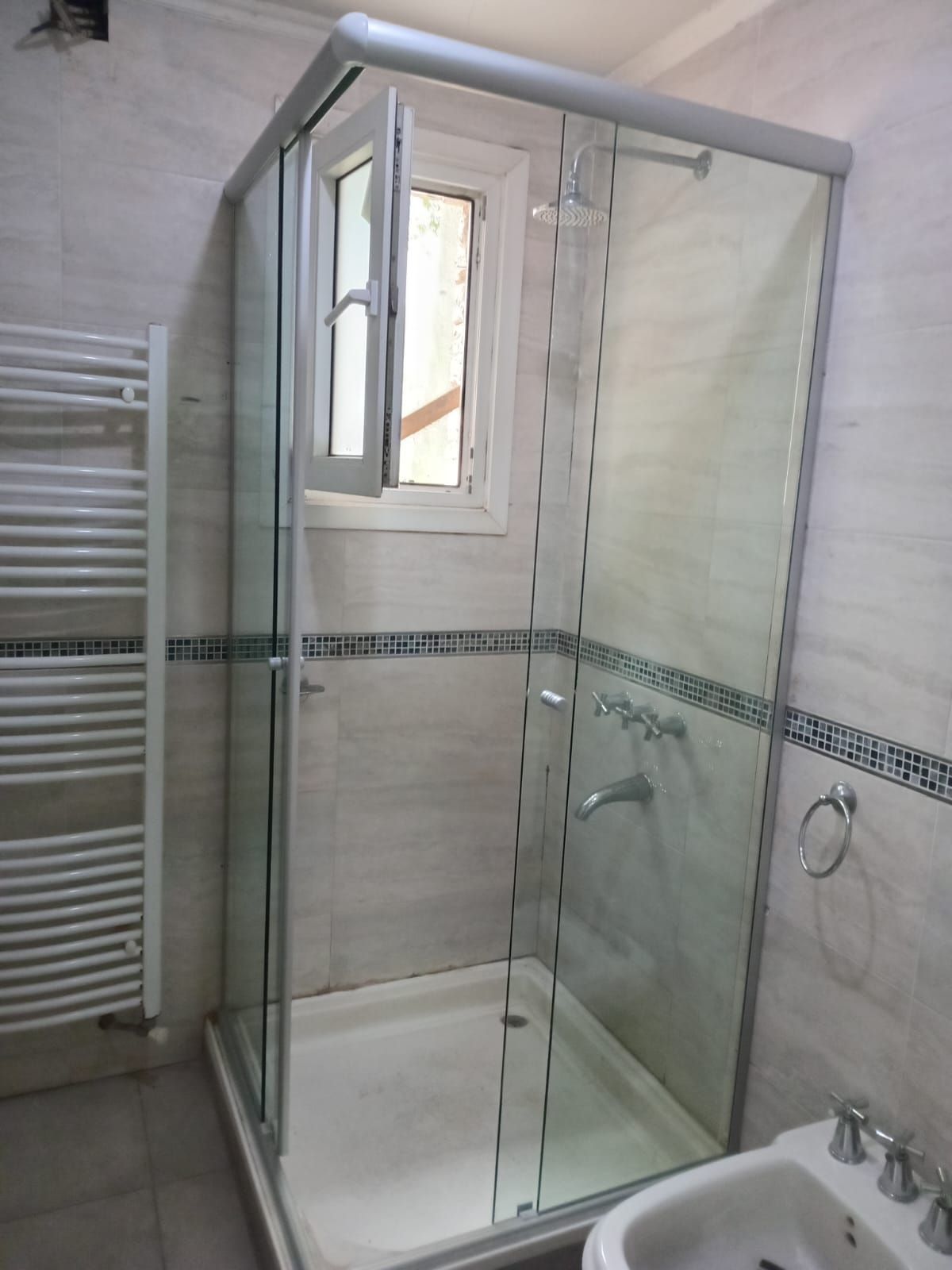 Cabina de ducha con puertas de cristal y ventana en un baño. Paredes blancas y grifería cromada.