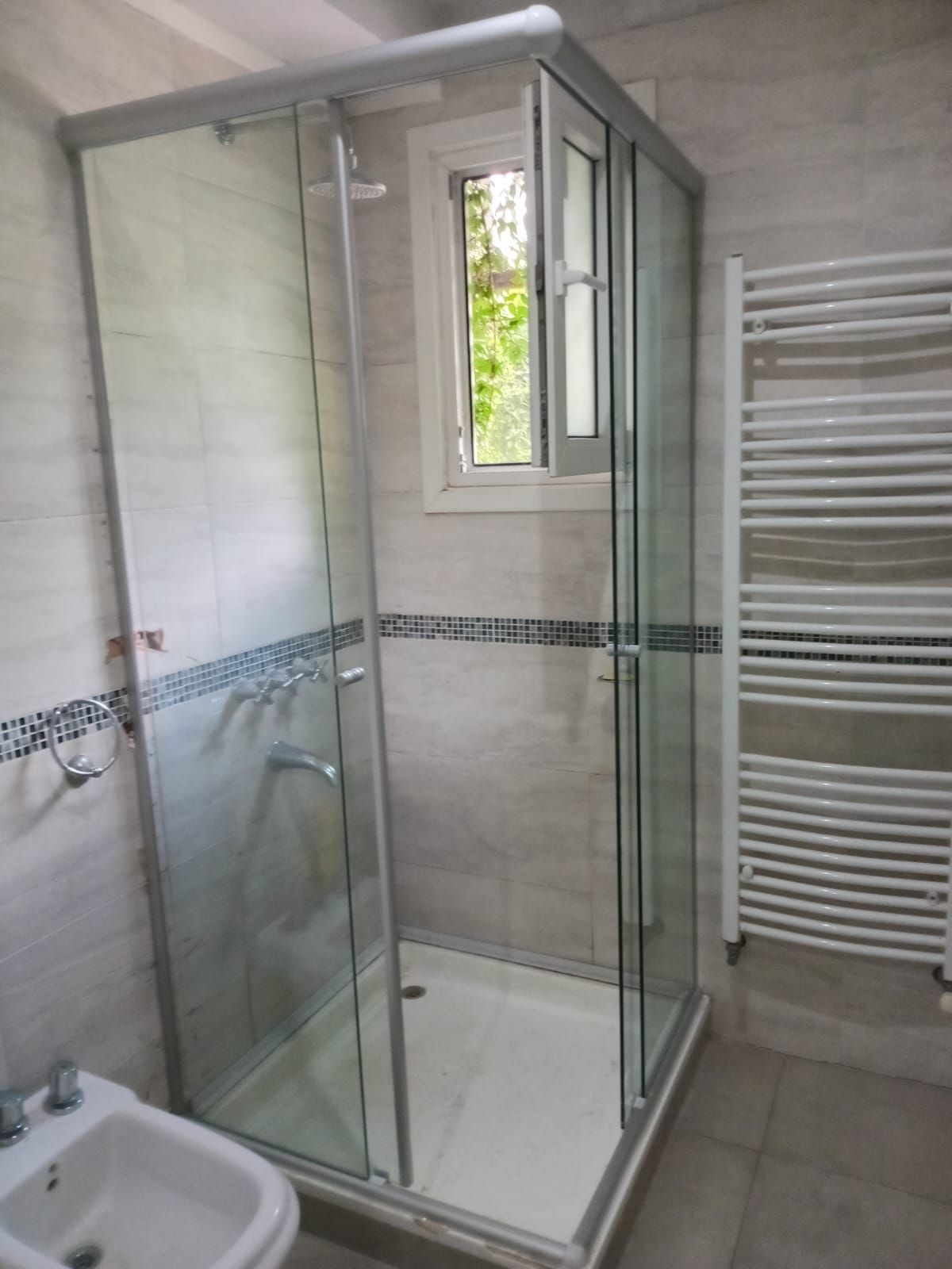 Cabina de ducha con puertas de cristal, paredes y molduras blancas, ventana pequeña y radiador.