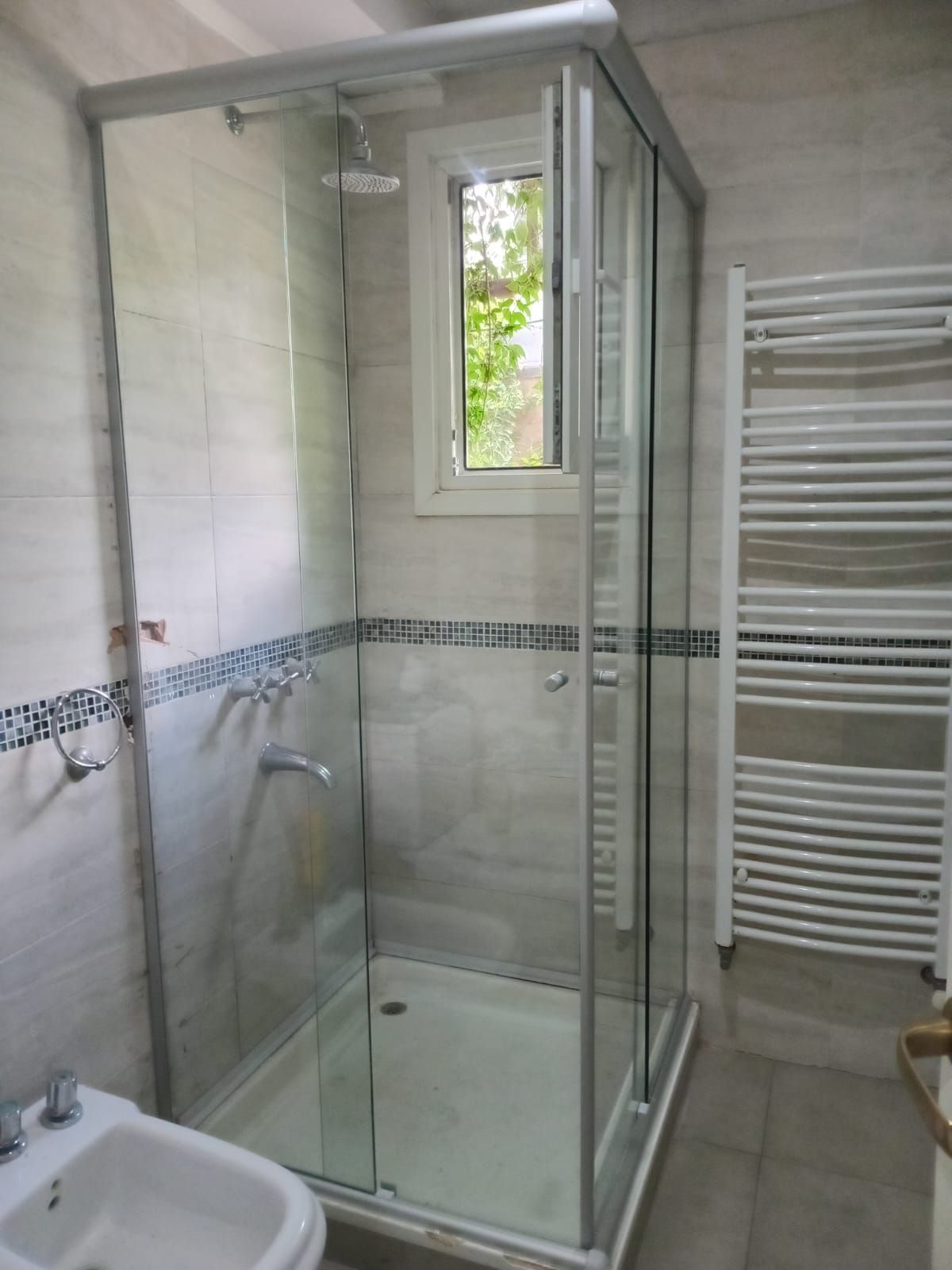 Cabina de ducha en un baño con puertas de cristal, ventana y toallero blanco.