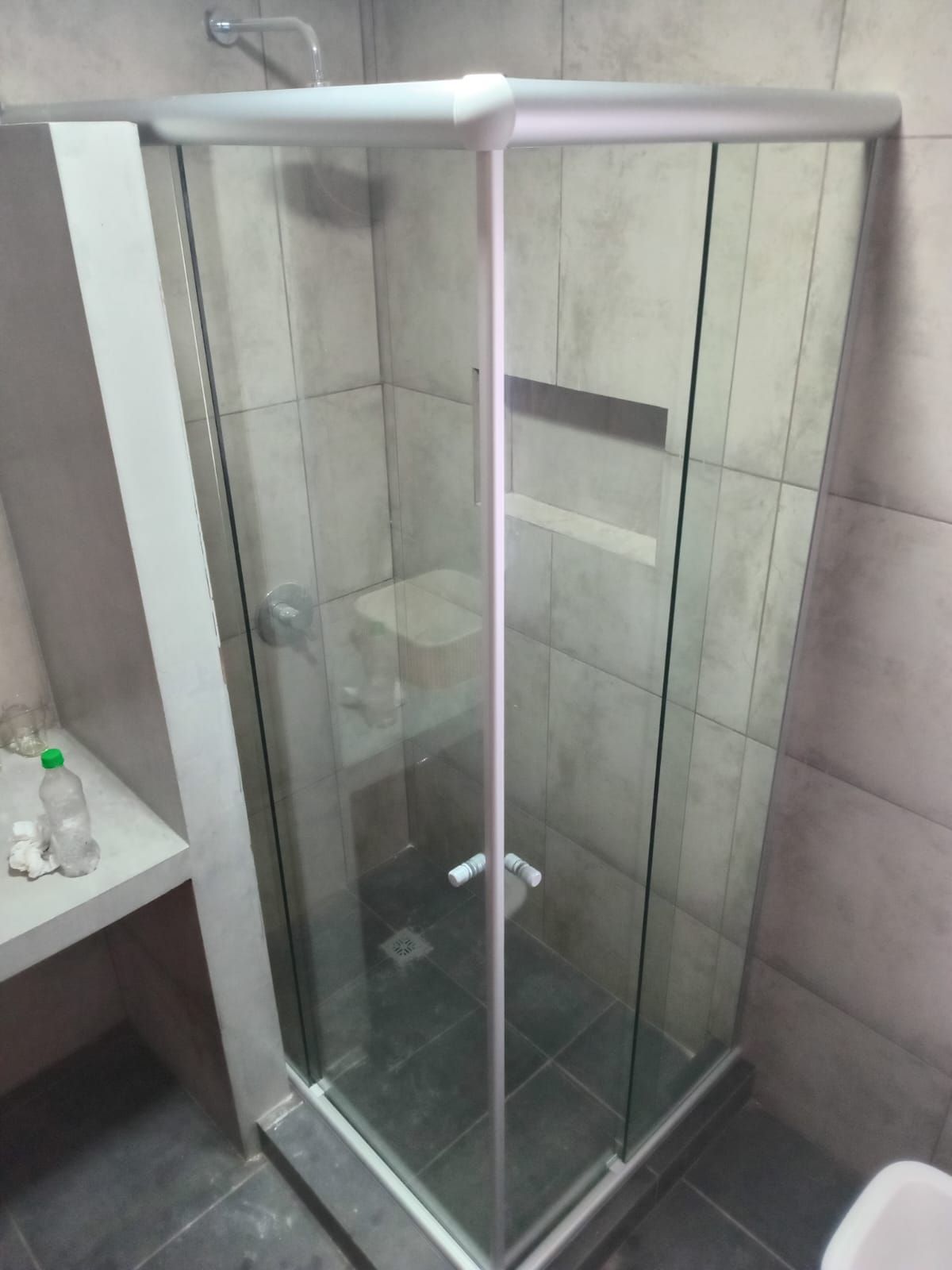 Una mampara de ducha de cristal con marco plateado en un baño con paredes y suelo de azulejos grises.