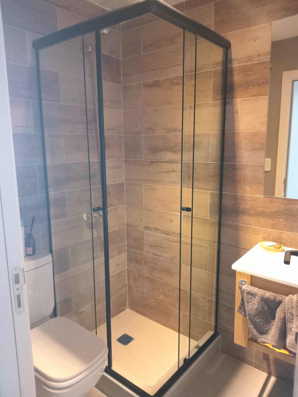 Cabina de ducha con marco negro y puertas de cristal transparente en un baño con paredes alicatadas y WC.