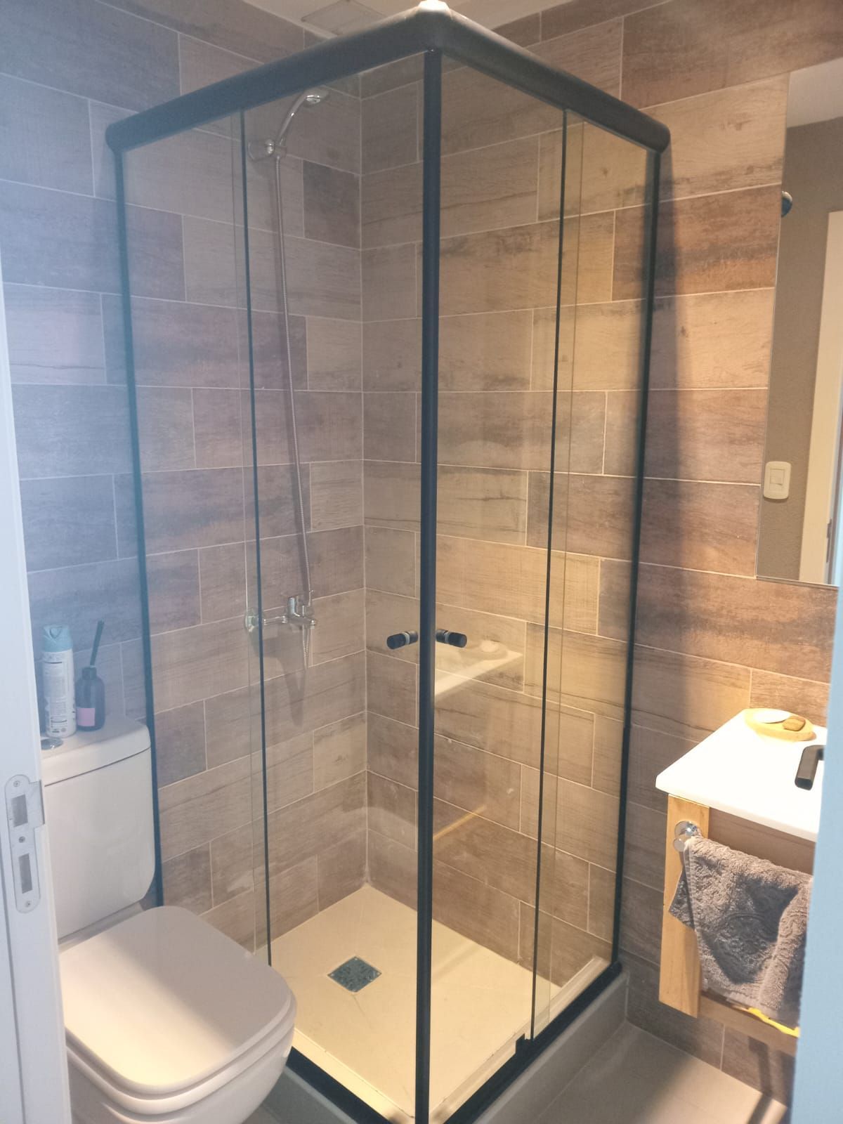 Cabina de ducha con marco negro, puertas de cristal y paredes de azulejos claros. Inodoro y lavabo visibles.