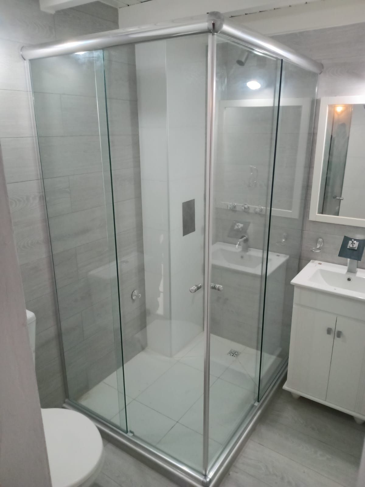 Baño con mampara de ducha de cristal, lavabo blanco, inodoro y paredes y suelo de azulejos grises.