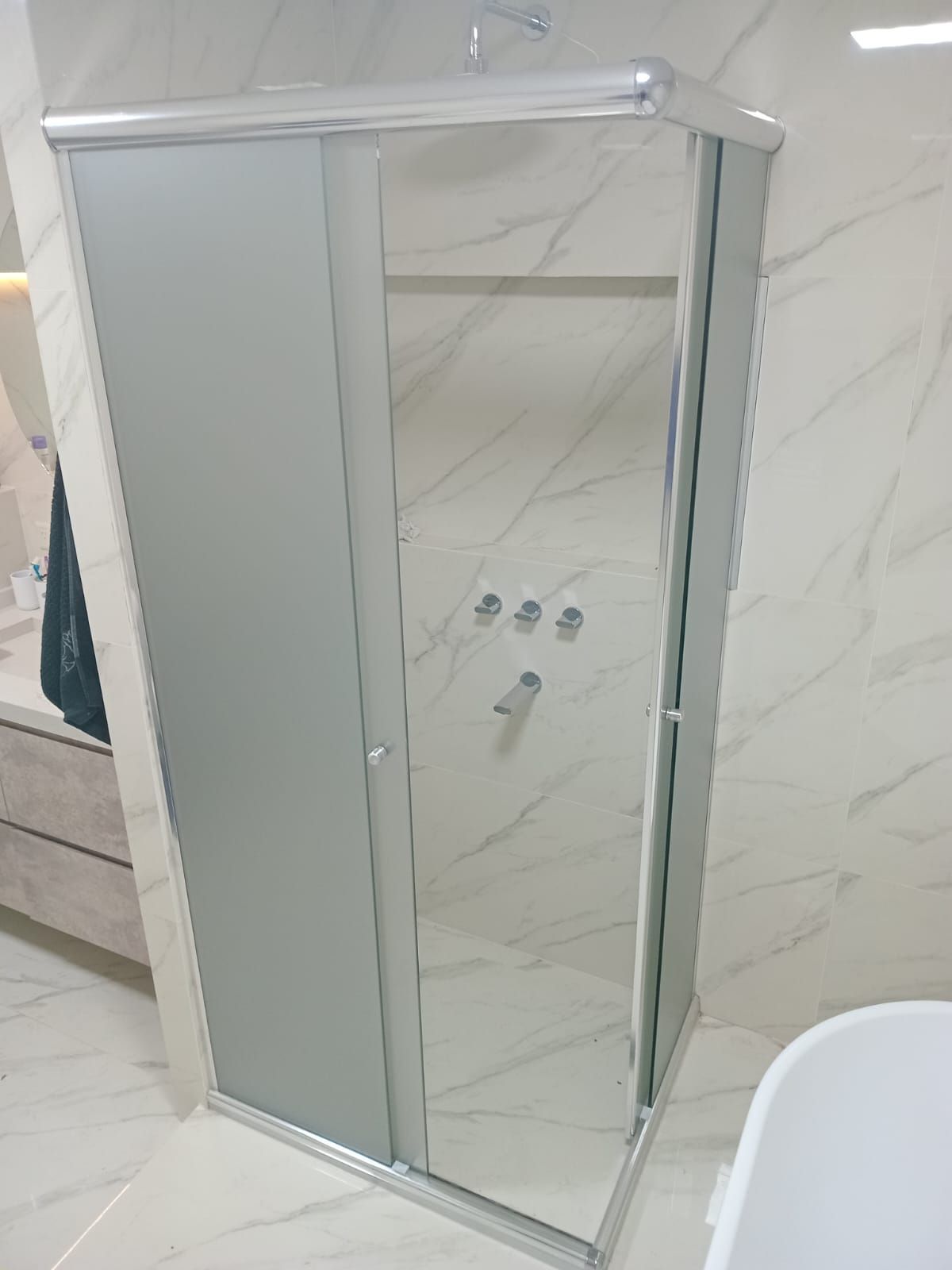 Mampara de ducha con paneles de cristal esmerilado y transparente, marco color plata, en baño revestido de azulejos blancos.