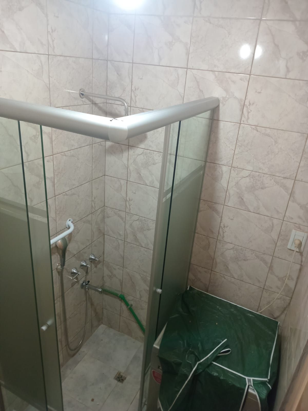Cabina de ducha con puertas de cristal y paredes de azulejos. Una lona verde cubre un objeto en la esquina.