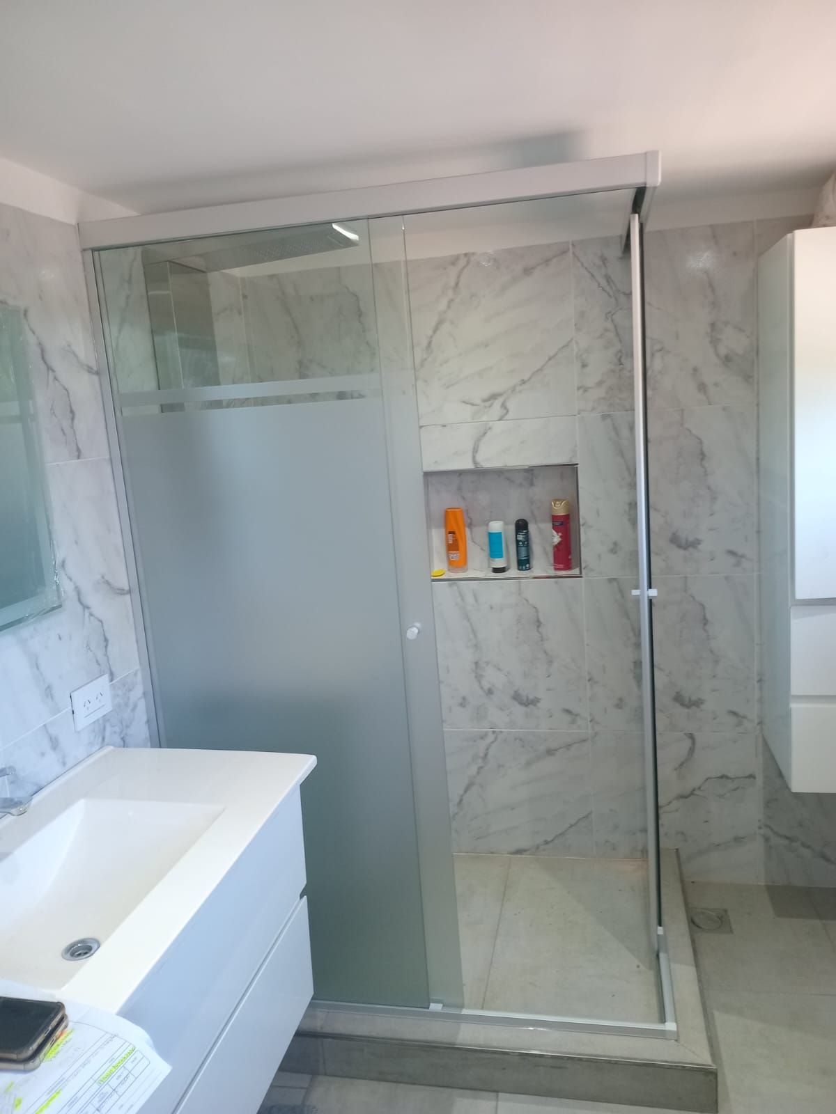 Baño con ducha de cristal, lavabo blanco y azulejos imitación mármol. El nicho de la ducha alberga artículos de aseo.