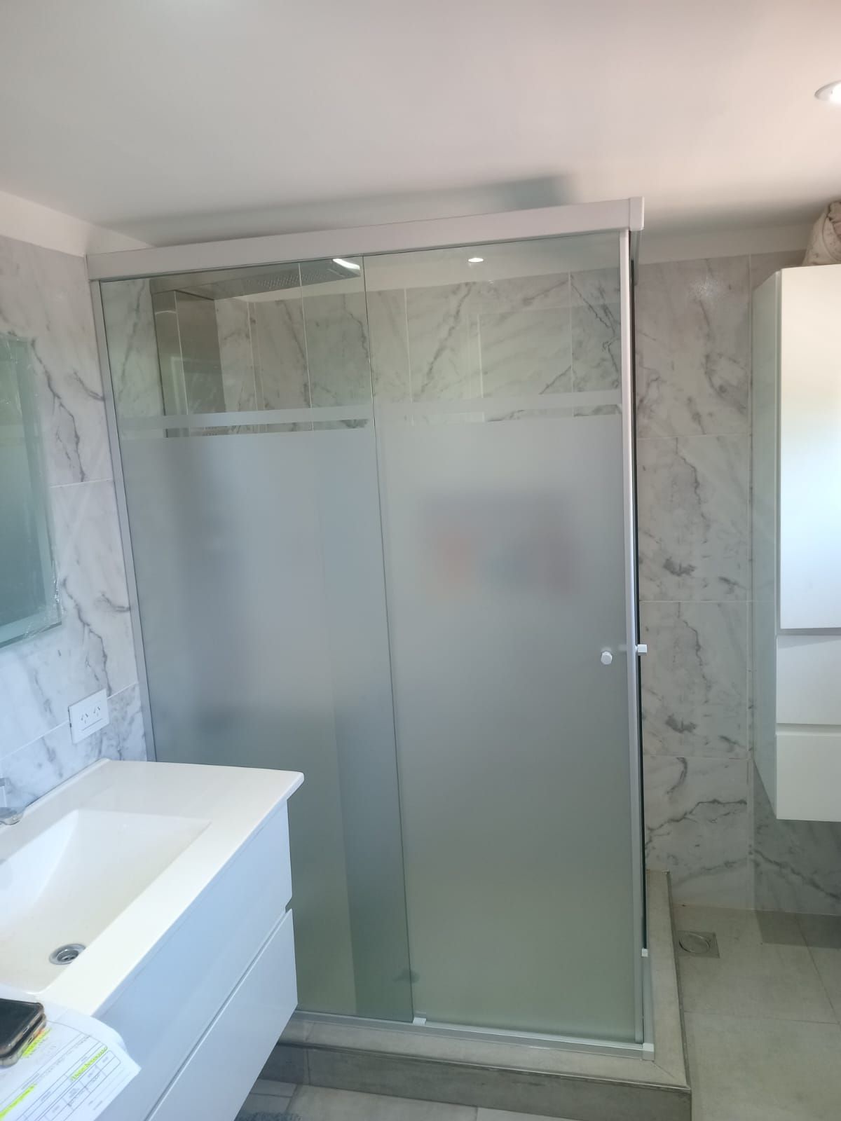 Baño con cabina de ducha, puerta de vidrio parcialmente esmerilado y tocador blanco.