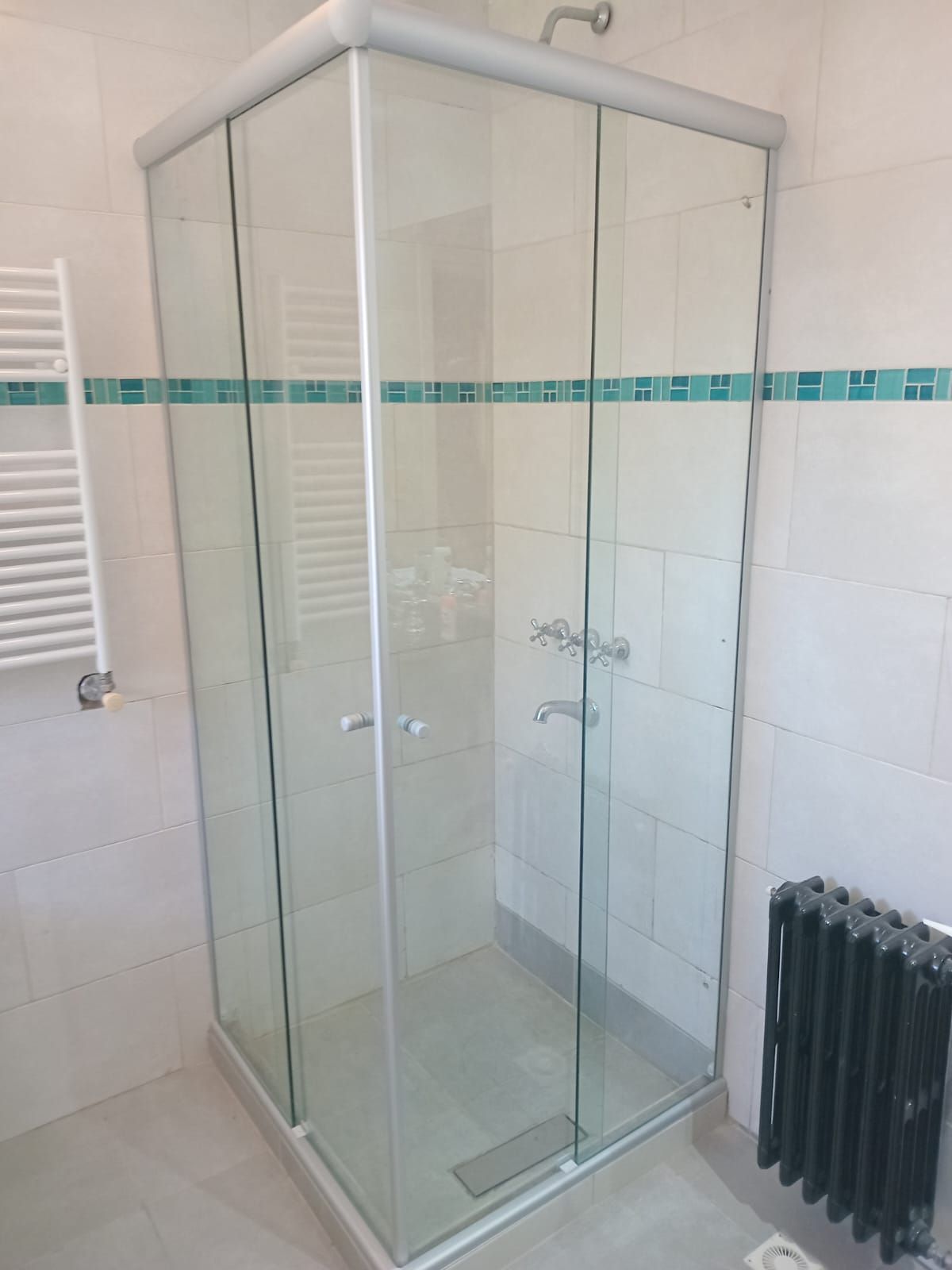 Mampara de ducha de cristal en un baño con azulejos blancos y detalles de mosaico color verde azulado.