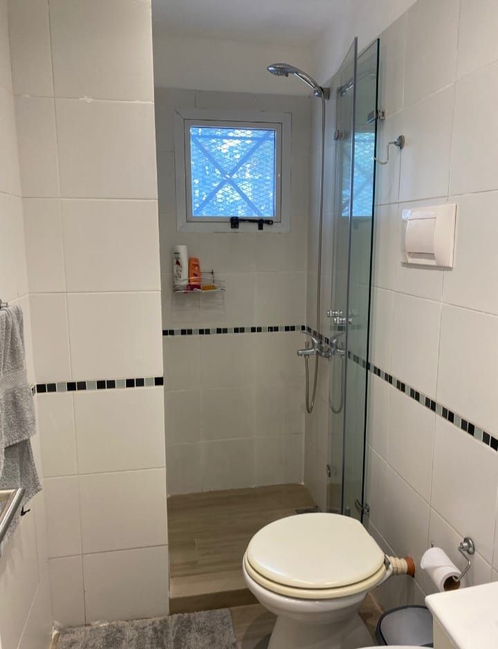 Baño con paredes de azulejos blancos, ducha, inodoro y pequeña ventana.