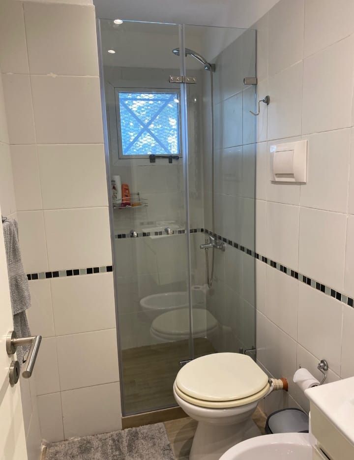 Baño con ducha de cristal, inodoro y ventana con vistas. Azulejos blancos y detalle de rayas negras.