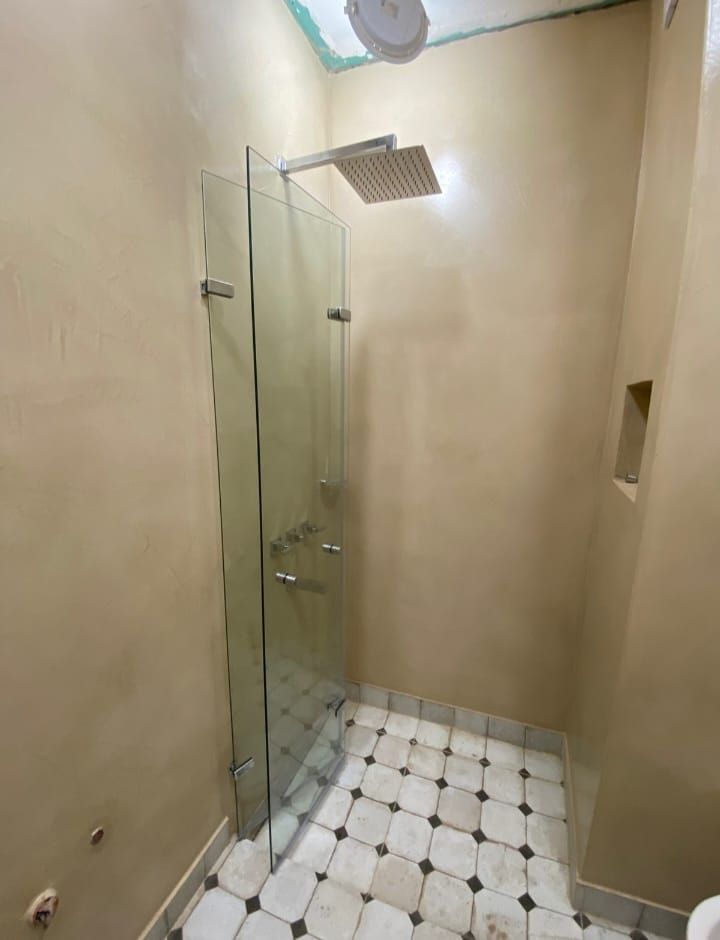 Cabina de ducha con puertas de vidrio, cabezal de ducha cuadrado, paredes de color beige y piso de baldosas estampadas.