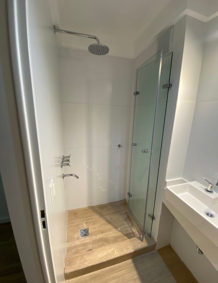 Cabina de ducha moderna con puerta de cristal, cabezal de ducha tipo lluvia y suelo imitación madera.