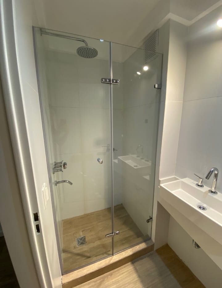 Mampara de ducha de cristal con cabezal de ducha tipo lluvia, junto a un lavabo blanco.