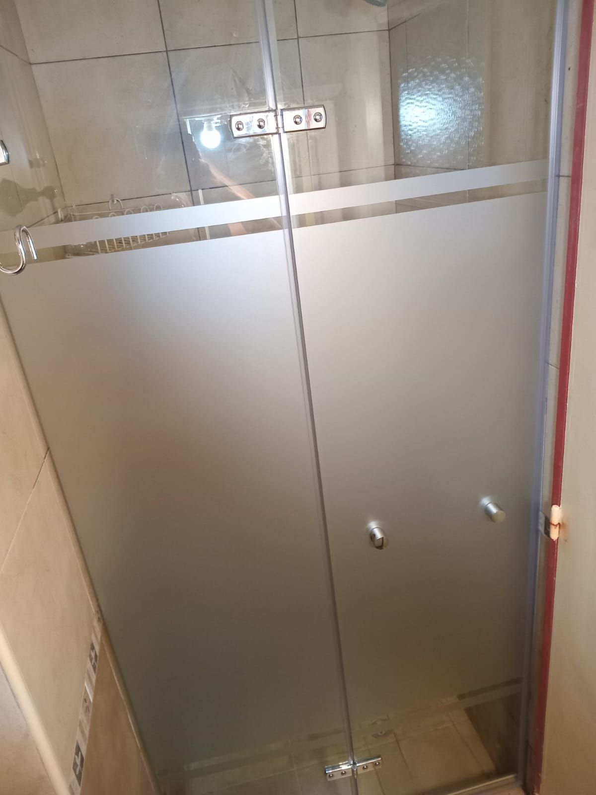 Puerta de ducha de vidrio con mitad inferior esmerilada, bisagras y manijas cromadas, en un baño con azulejos de color beige.