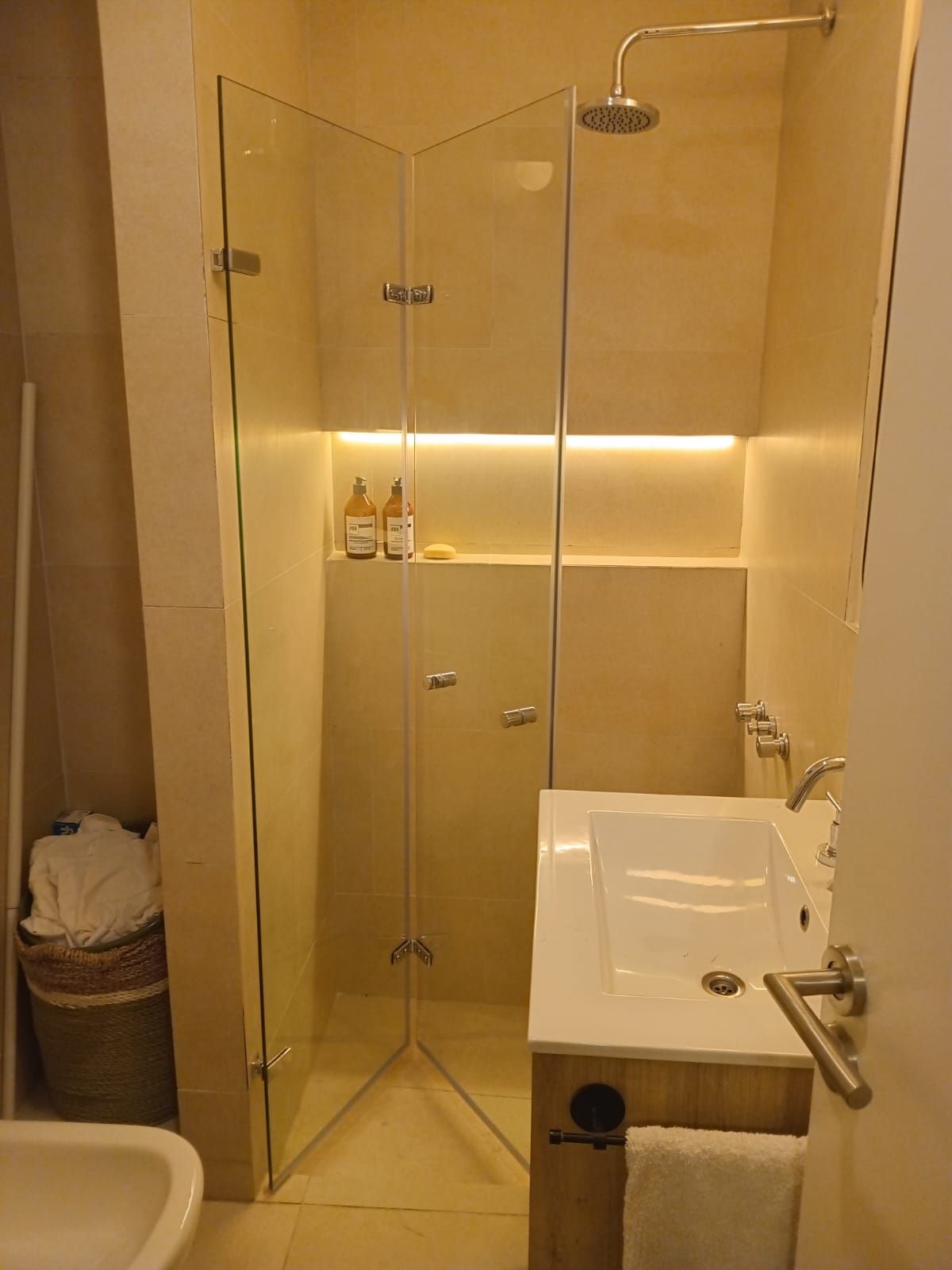 Baño con ducha de esquina, lavabo y cesto para ropa.