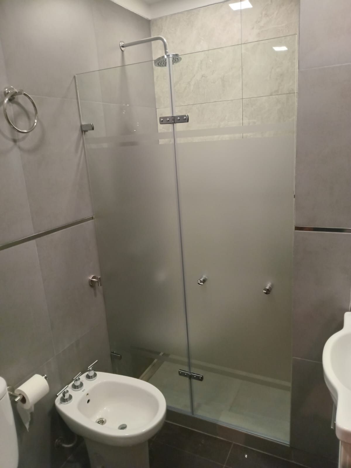Baño con mampara de ducha de cristal y bidé. También se aprecian paredes y azulejos grises, y un inodoro.