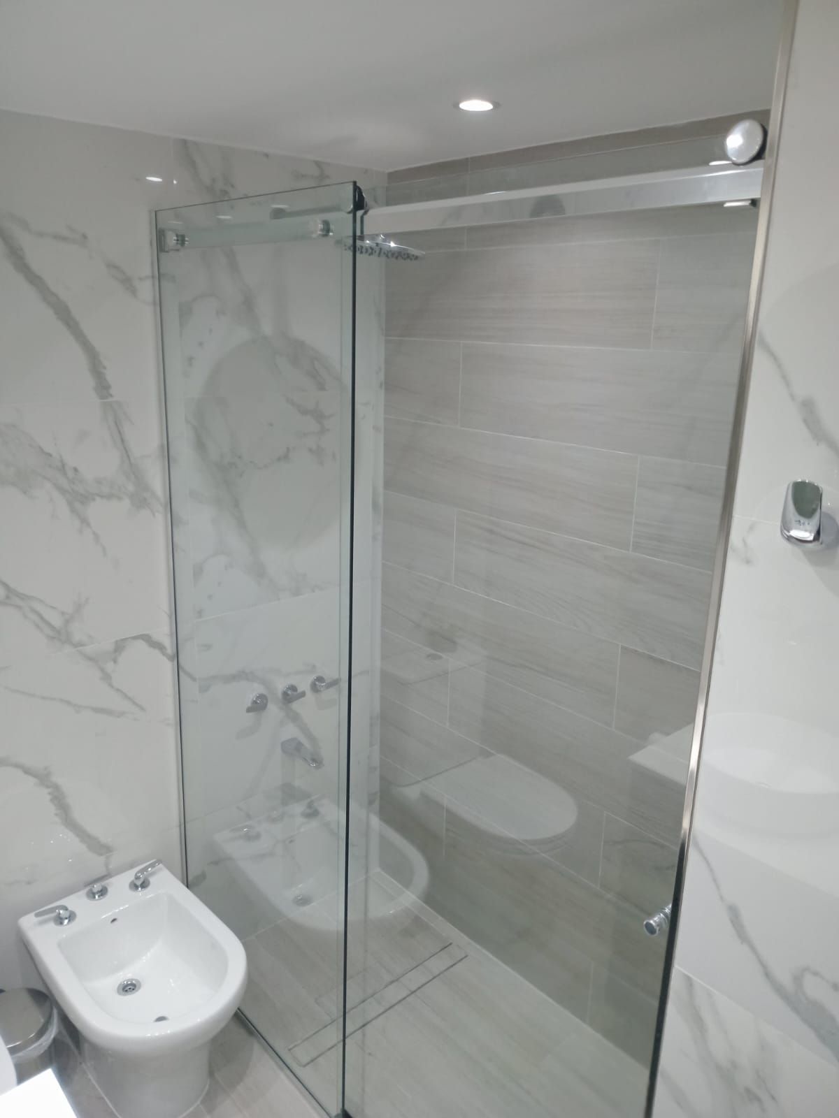 Baño moderno con mampara de ducha de cristal, bidé e inodoro. Paredes de azulejos blancos y grises.