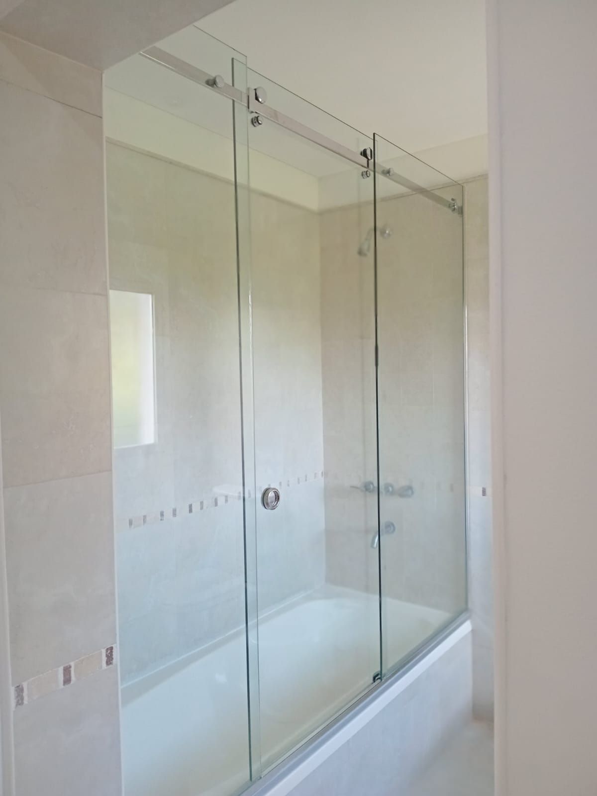 Mampara de ducha de cristal sobre bañera, con puerta corredera y herrajes plateados, en baño de azulejos blancos.