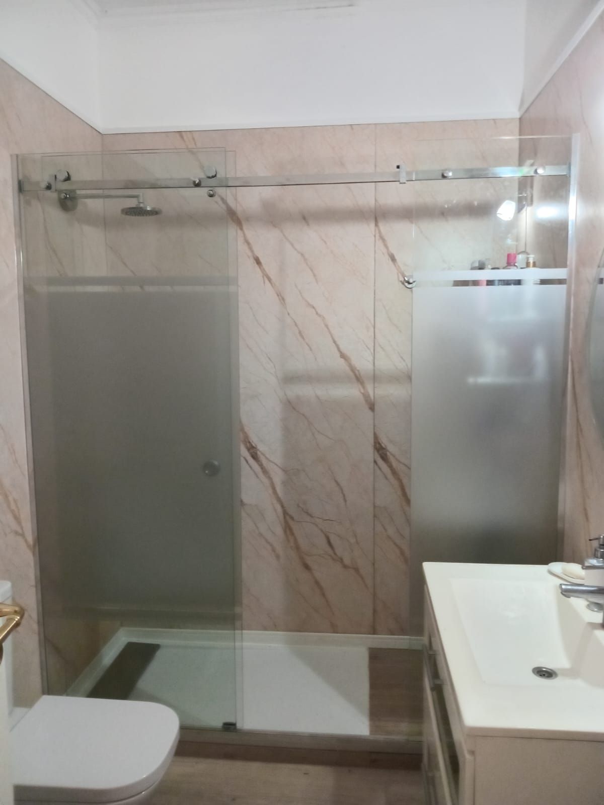Baño con ducha con mampara corredera de cristal y lavabo blanco. Paredes con diseño de mármol.