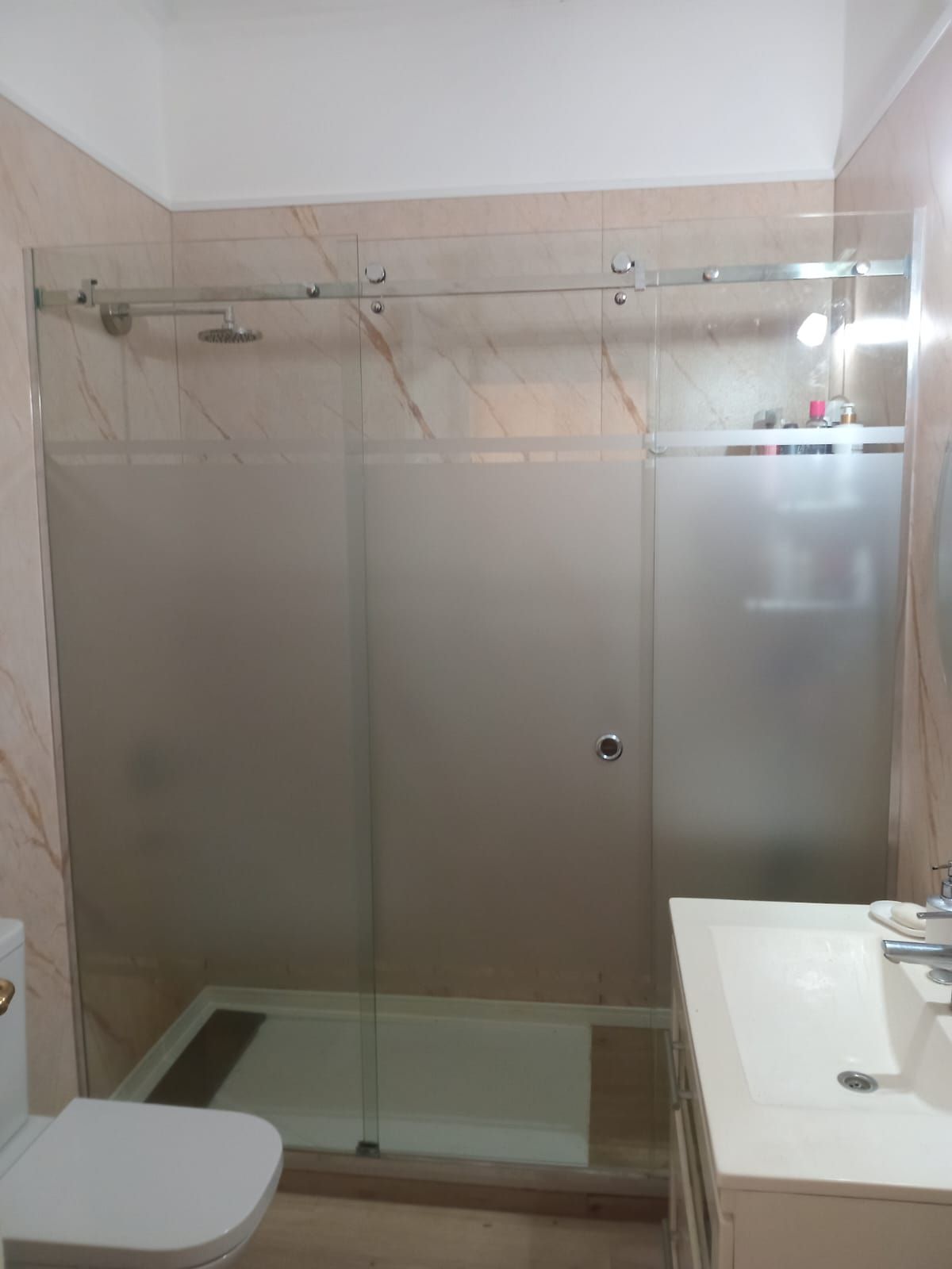 Ducha con puertas de vidrio esmerilado, paredes de azulejos de color beige claro, lavabo e inodoro blancos.