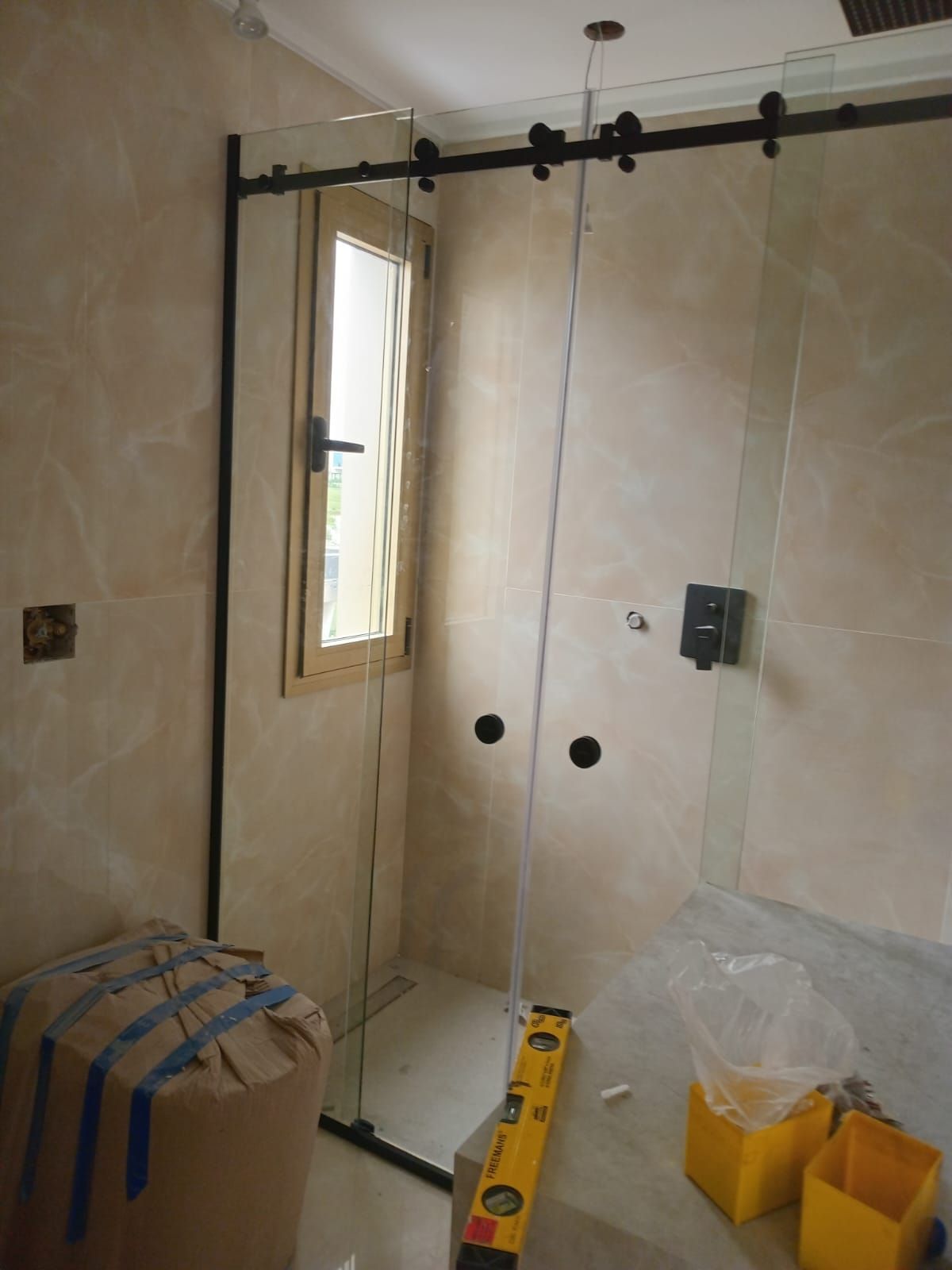 Cabina de ducha en construcción, cuenta con herrajes negros, vidrio transparente y paredes de azulejos de color claro.