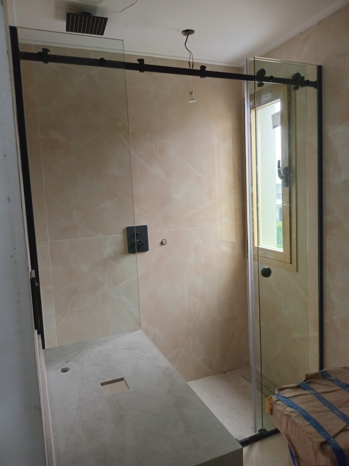 Cabina de ducha con puerta corredera de cristal y marco negro. La cabina tiene azulejos beige y suelo de hormigón claro.