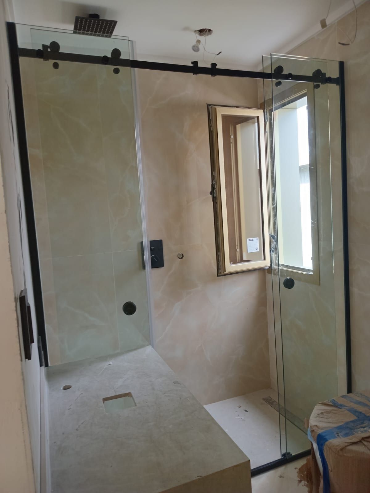 Cabina de ducha con puertas de cristal, marco negro y una pequeña ventana. Sin terminar, en tonos neutros.